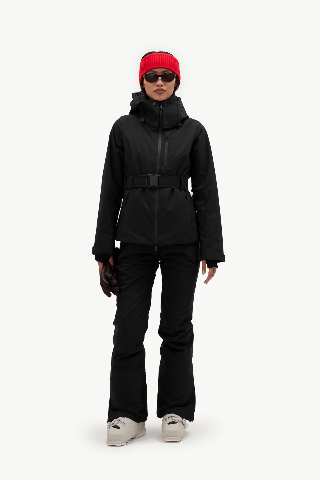 A model wearing a black hooded ski jacket with a matching black belt, black pants, a bright red knit beanie, and black sunglasses, shown against a plain white studio background.