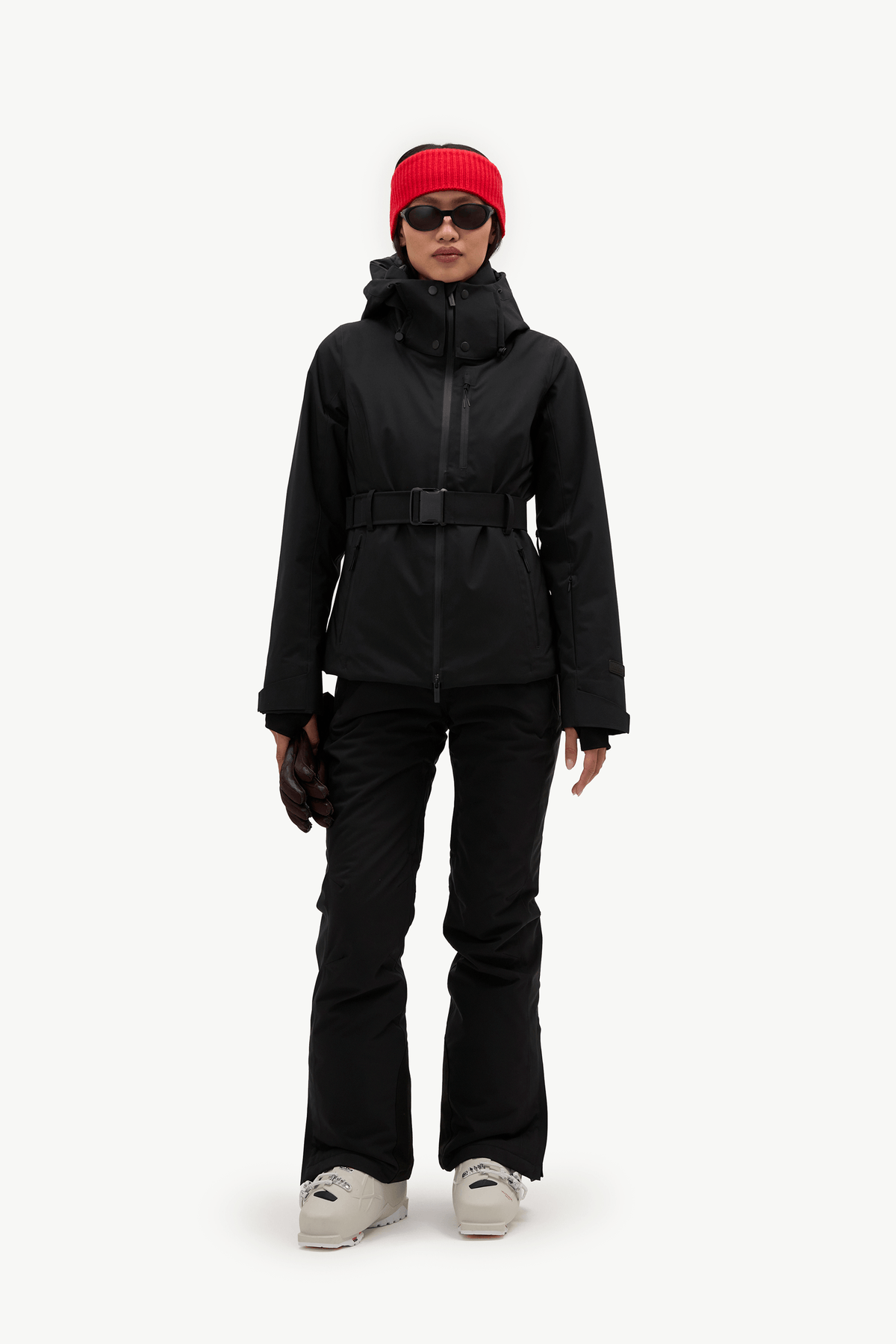 A model wearing a black hooded ski jacket with a matching black belt, black pants, a bright red knit beanie, and black sunglasses, shown against a plain white studio background.