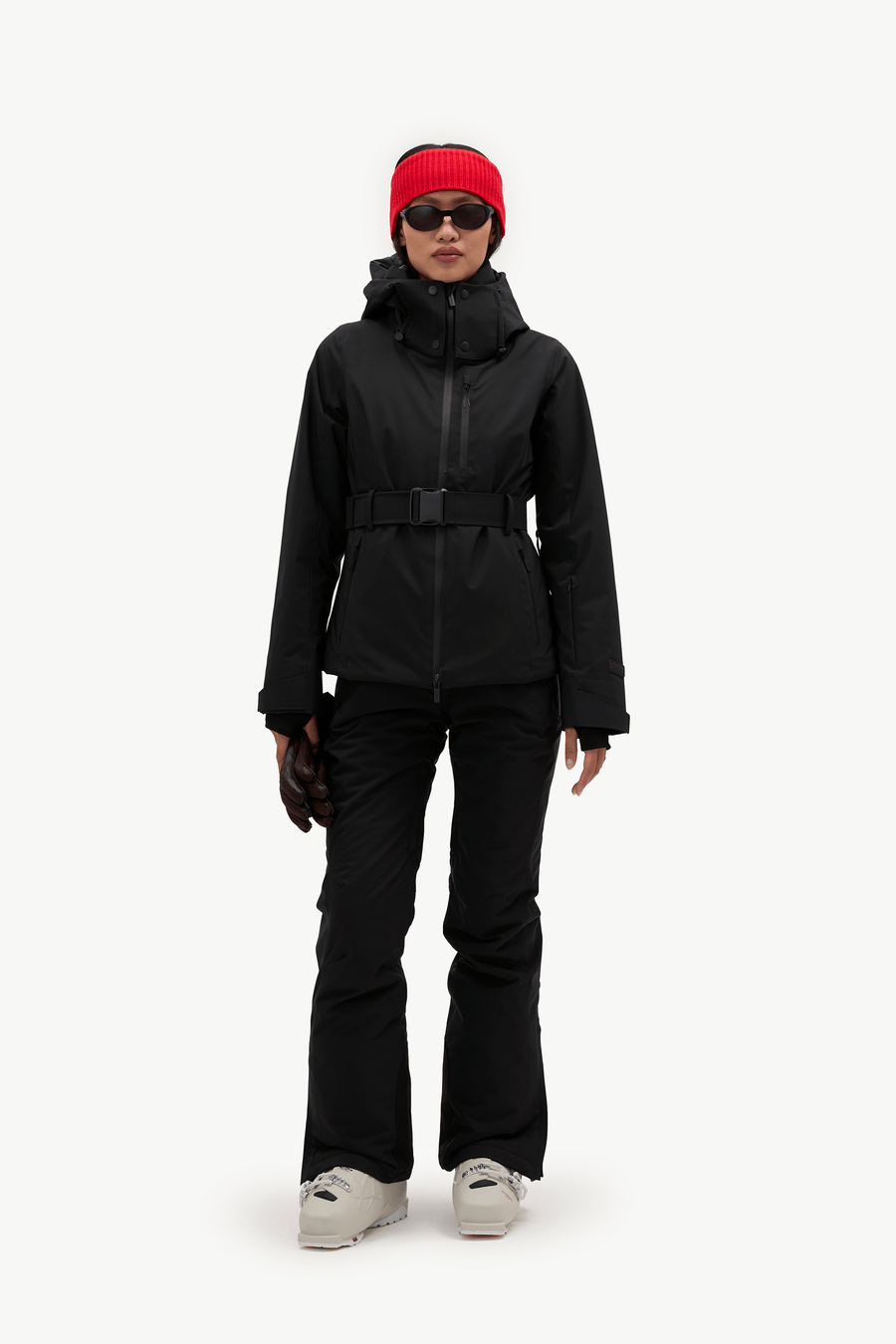 A model wearing a black hooded ski jacket with a matching black belt, black pants, a bright red knit beanie, and black sunglasses, shown against a plain white studio background.
