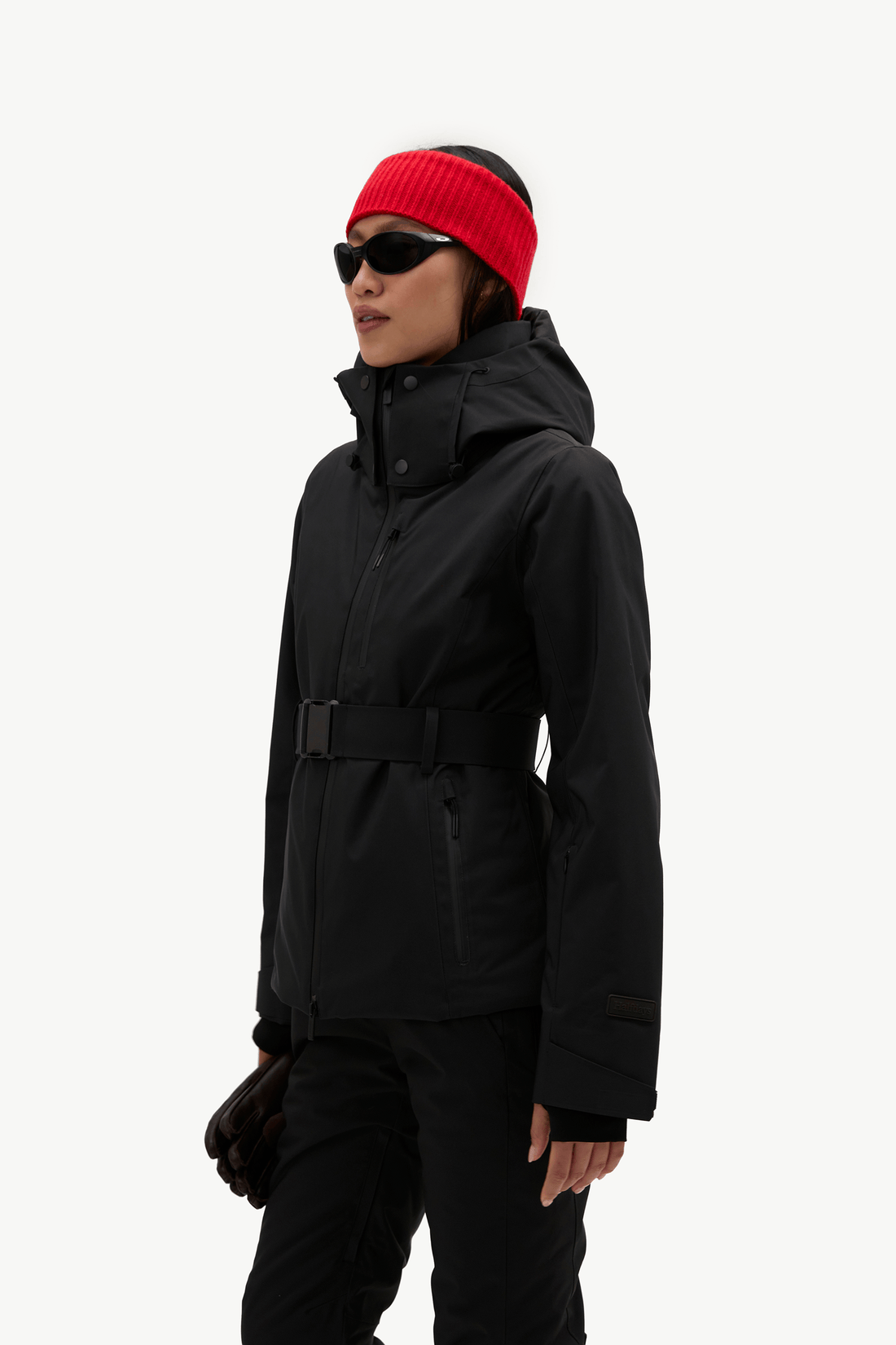 A model wearing a black hooded ski jacket with a matching black belt, black pants, a bright red knit beanie, and black sunglasses, shown against a plain white studio background.