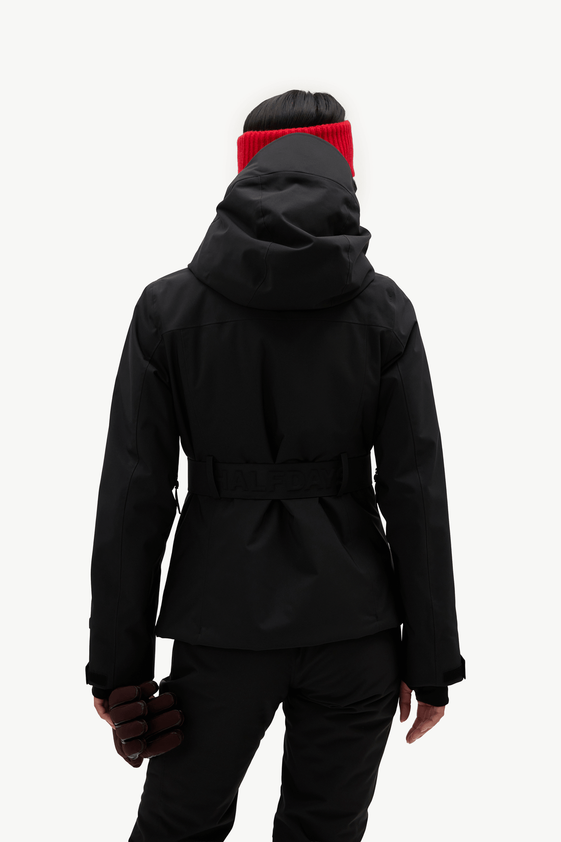 A model wearing a black hooded ski jacket with a matching black belt, black pants, a bright red knit beanie, shown against a plain white studio background.
