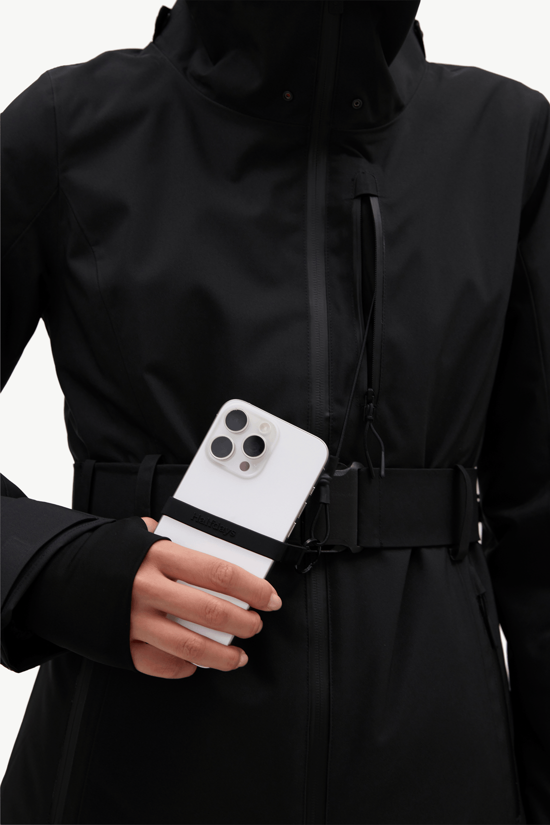 A close-up of a person wearing a black hooded jacket with a belt and side zipper pocket, holding a white smartphone in the pocket.