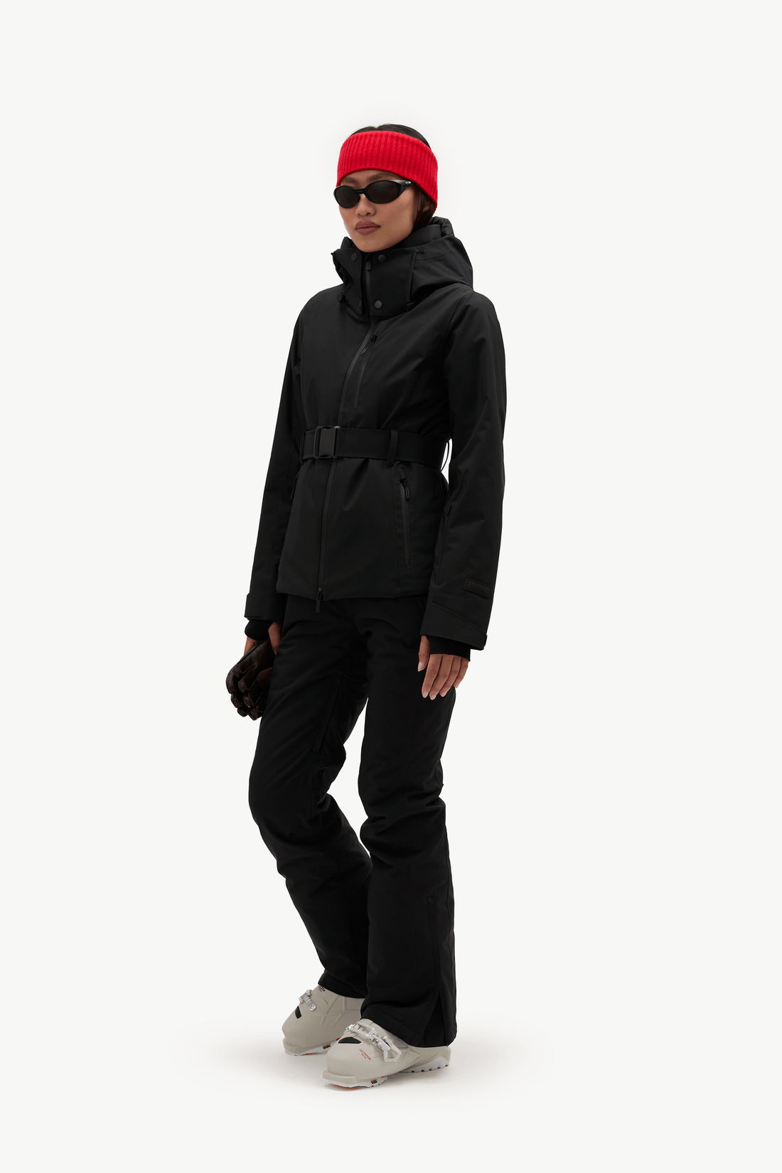 A model wearing a black ski jacket with a matching black ski pants, a bright red knit beanie, black sunglasses, and light gray snowboard boots, standing against a solid white background.