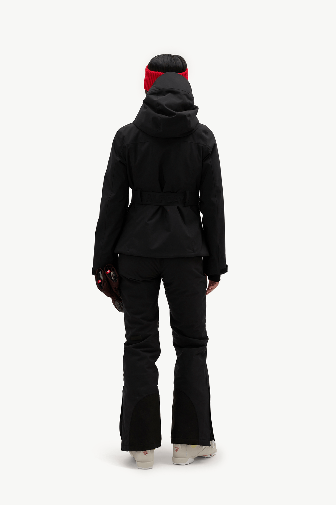 A model shown from the back wearing a black hooded jacket with a waist belt, matching black flared ski pants, a black beanie with a red band, and white sneakers, set against a plain white studio background.
