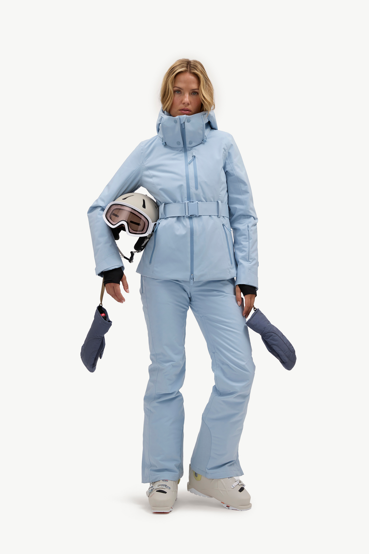 A woman modeling a light blue ski jacket and matching ski pants, holding a white ski helmet with goggles and dark blue gloves against a plain white studio background.