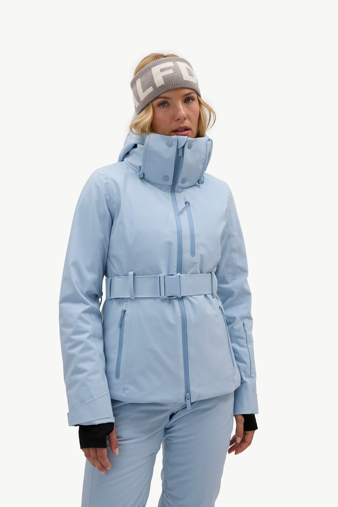 A woman modeling a light blue ski jacket with a matching belt and pants, wearing a gray beanie with white lettering, against a plain white studio background.
