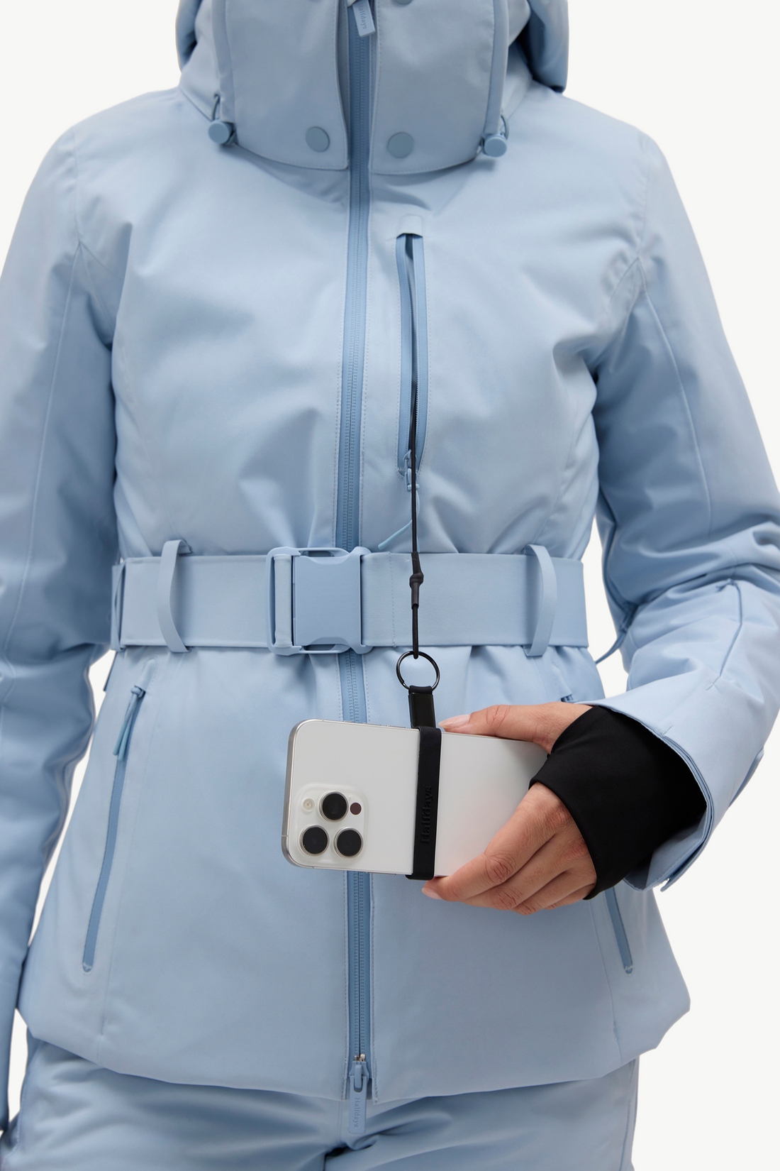 A woman modeling a light blue ski jacket and matching ski pants, holding a phone against a plain white studio background.