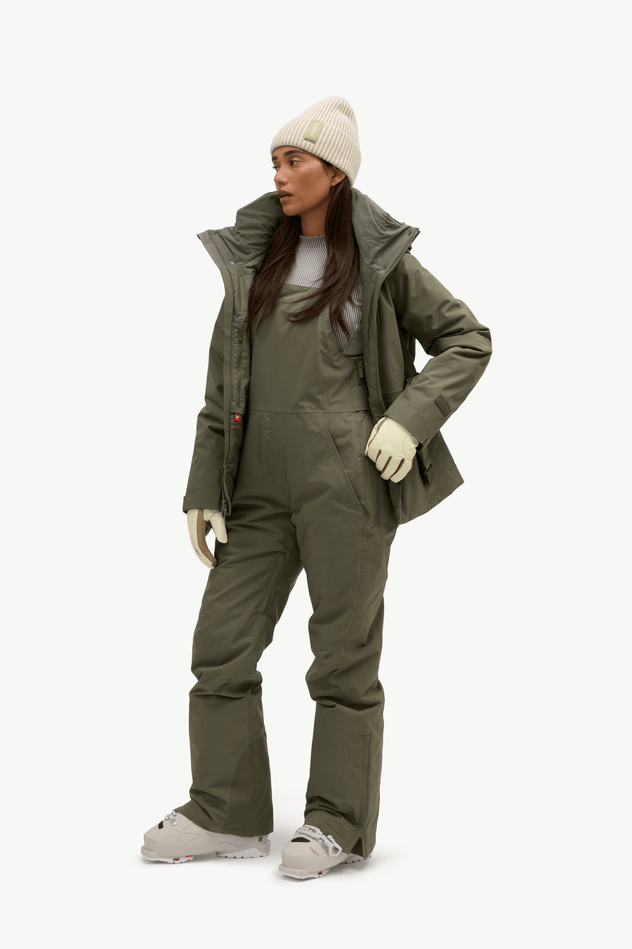 A woman modeling an olive green ski jacket and matching insulated bib pants, paired with a cream beanie, cream gloves, and light gray ski boots, against a plain white studio background.