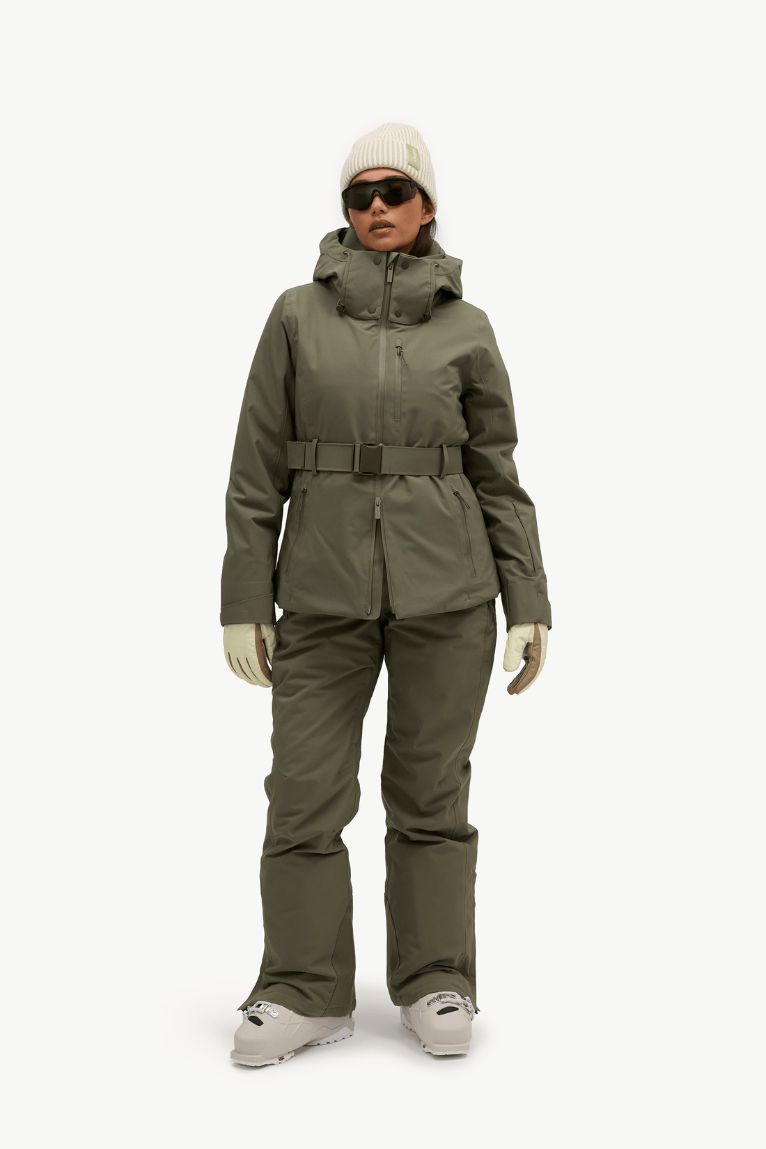 A model wearing a matching olive green ski jacket and pants set with a matching beanie, gloves, and snow boots, standing against a plain white studio background.