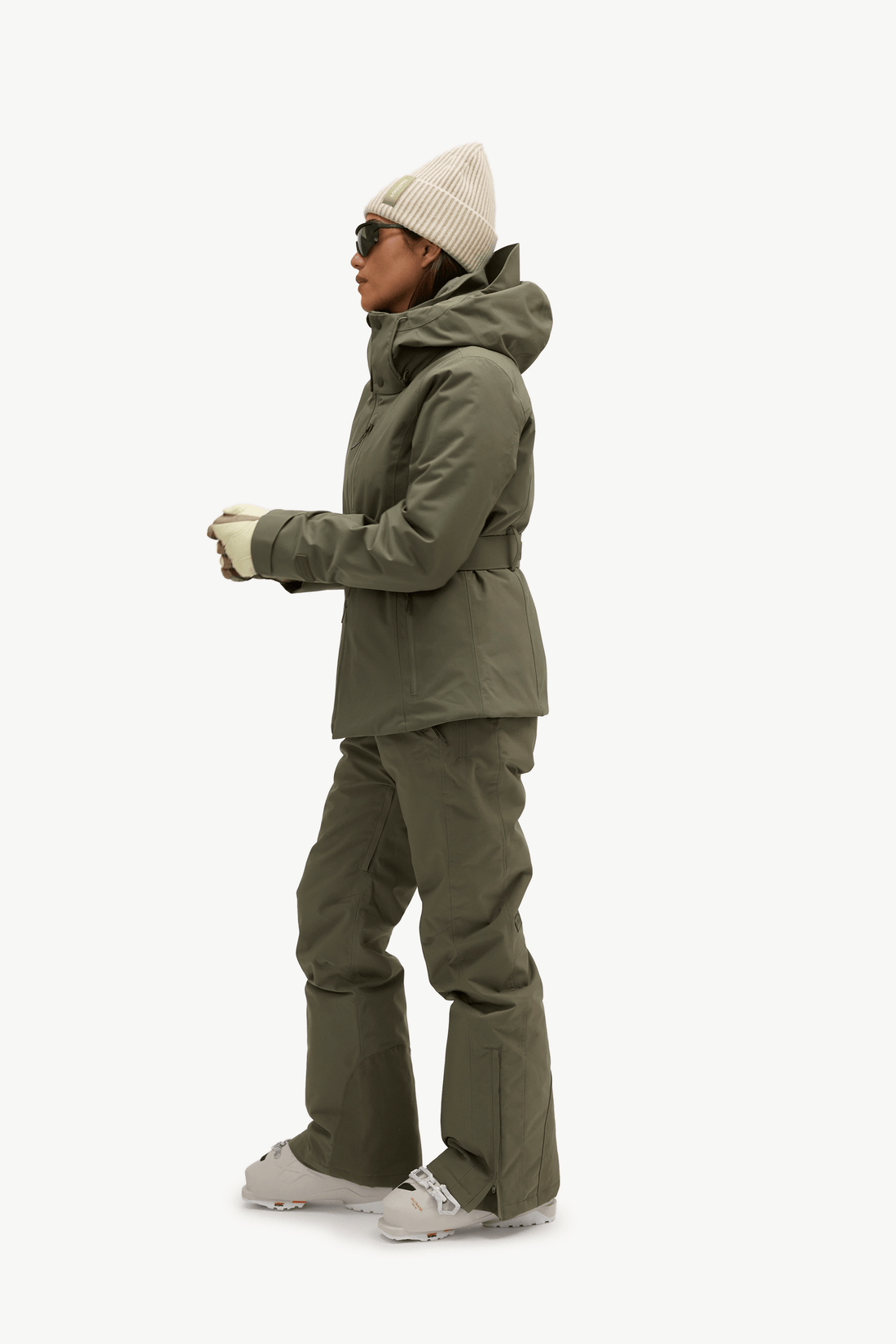 A model shown in profile wearing a matching olive green snow suit, consisting of a hooded jacket and insulated pants, along with a cream knit beanie, gloves, and ski boots against a solid off-white background.