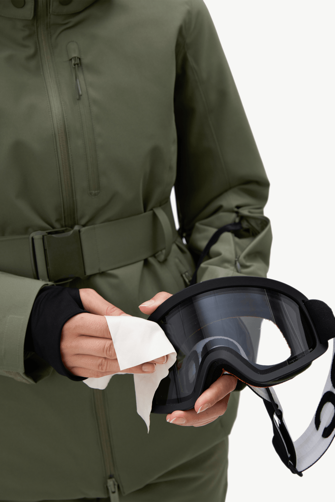 A close-up of a person wearing an olive green ski jacket and holding a pair of black ski goggles with a white lens protector while wiping them with a tissue.