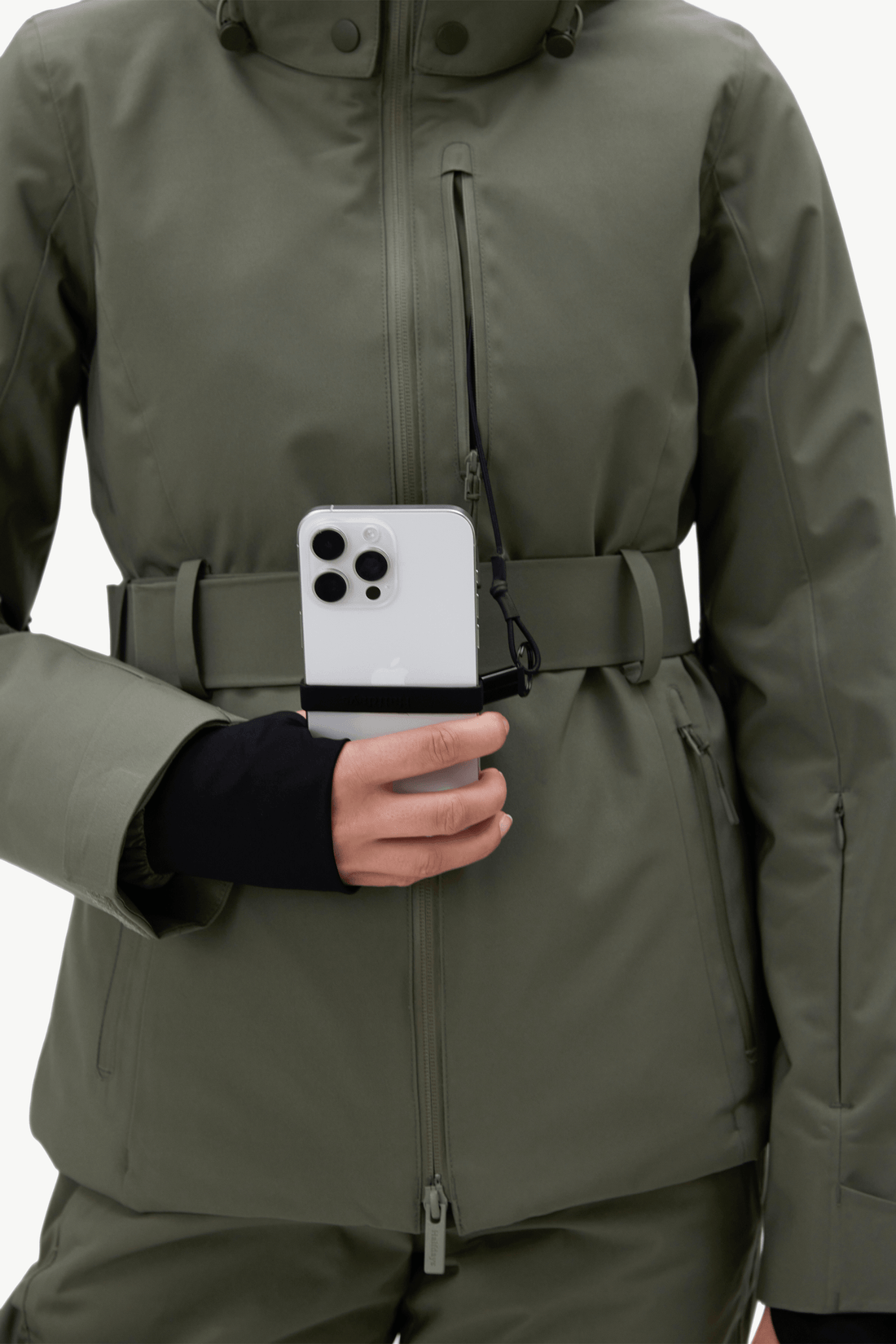 An olive green ski jacket with a matching belt and pants, featuring a unique front zipper pocket holding a white smartphone.