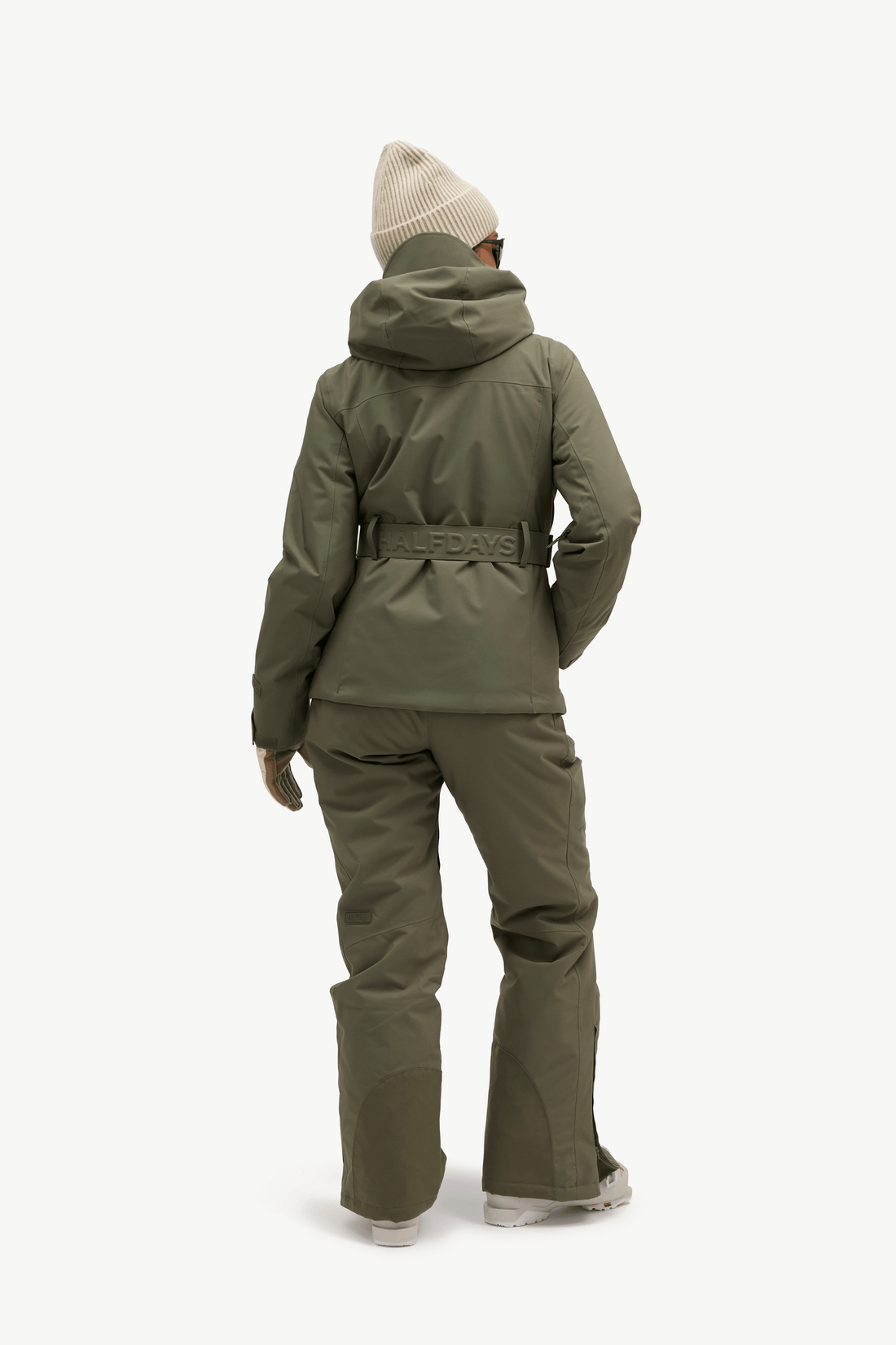 A model shown from the back wearing an olive green ski jacket and matching pants, paired with a cream knit beanie and white sneakers, against a plain white studio background.