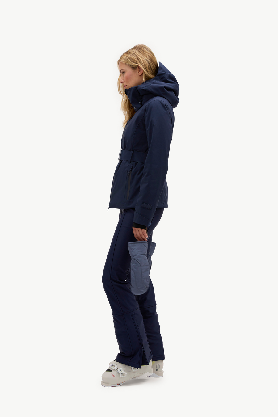 A woman in profile wearing a dark navy hooded jacket, matching navy pants, and light gray ski boots, standing against a plain white studio background.
