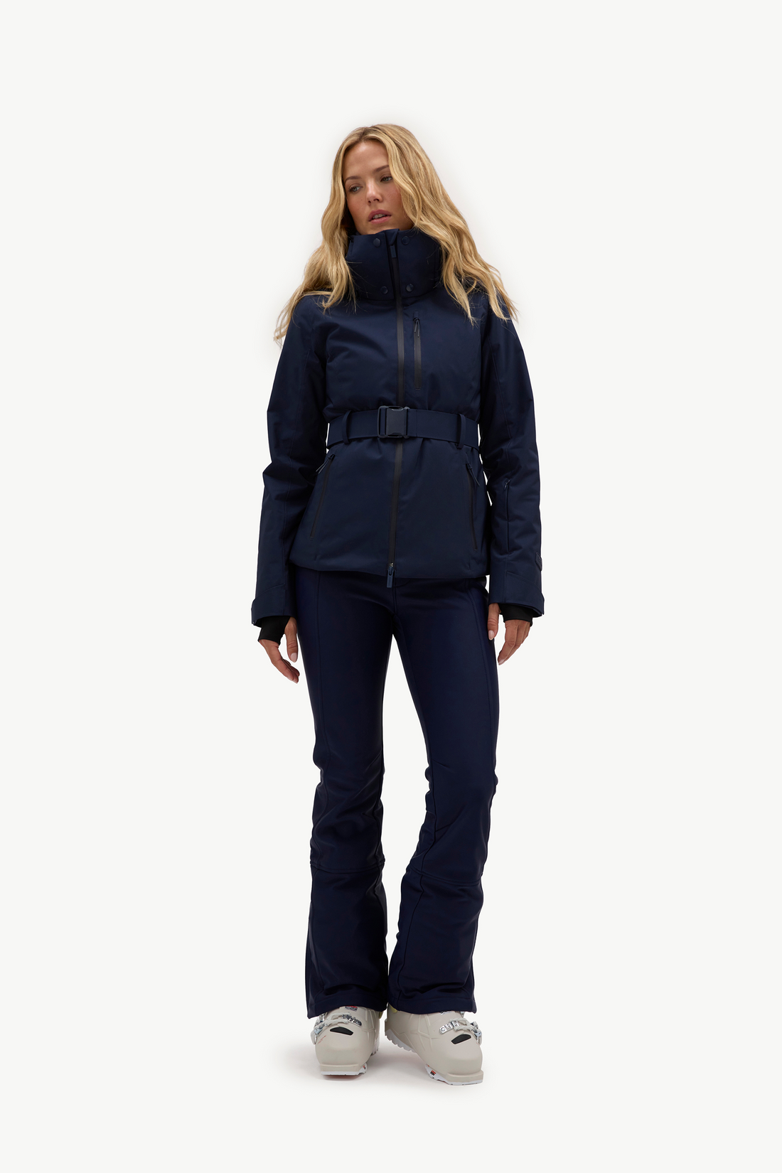 A woman wearing a dark navy hooded jacket, matching navy pants, and light gray ski boots, standing against a plain white studio background.