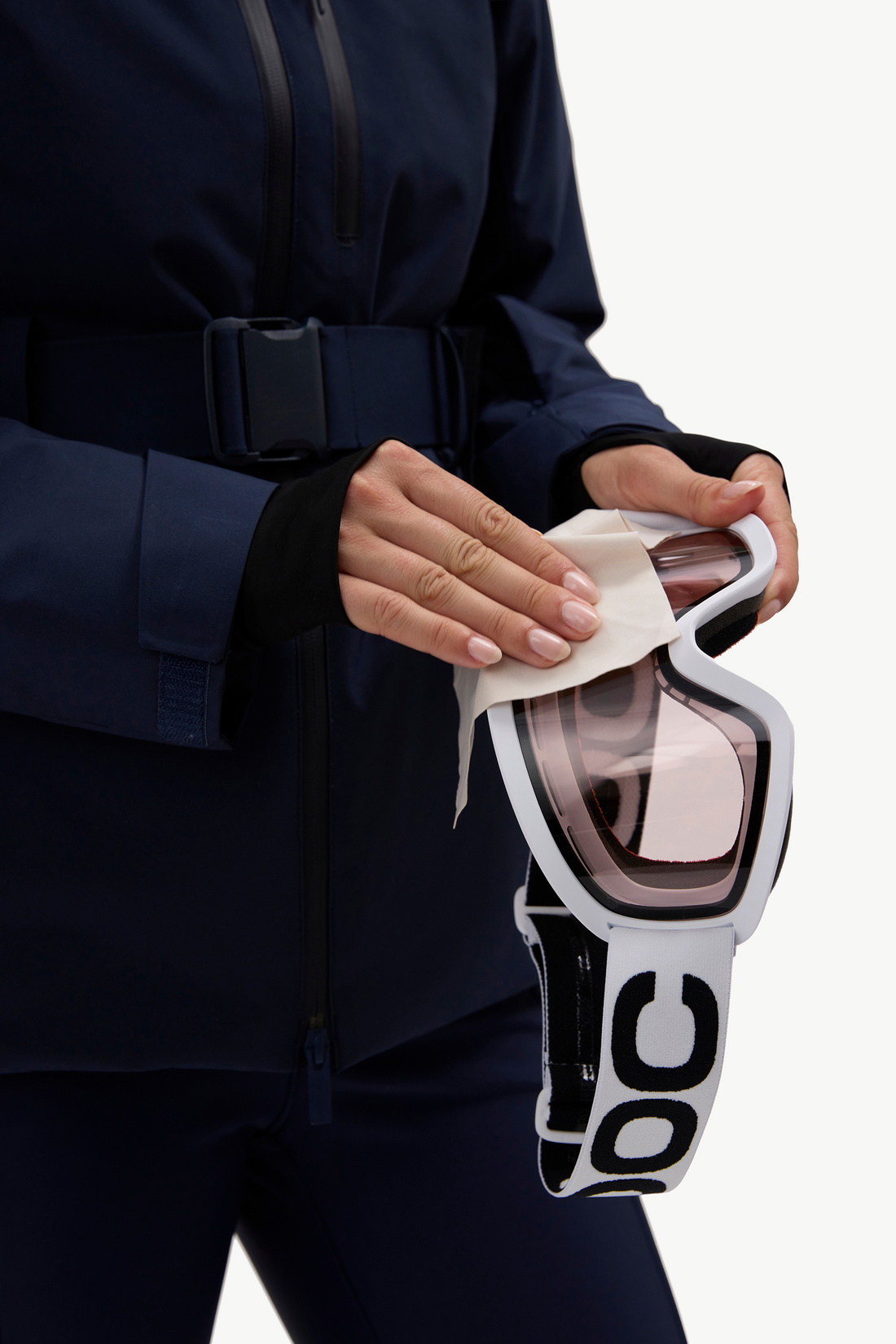A close-up of a person in a dark navy ski jacket holding a pair of white goggles with a reflective pink-tinted lens and a white strap, using a cloth to wipe the lens.