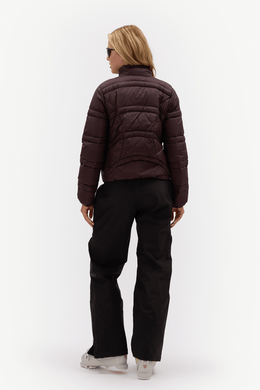 A woman viewed from behind wearing a dark plum-brown quilted puffer jacket, black pants, and white sneakers against a plain white studio background.