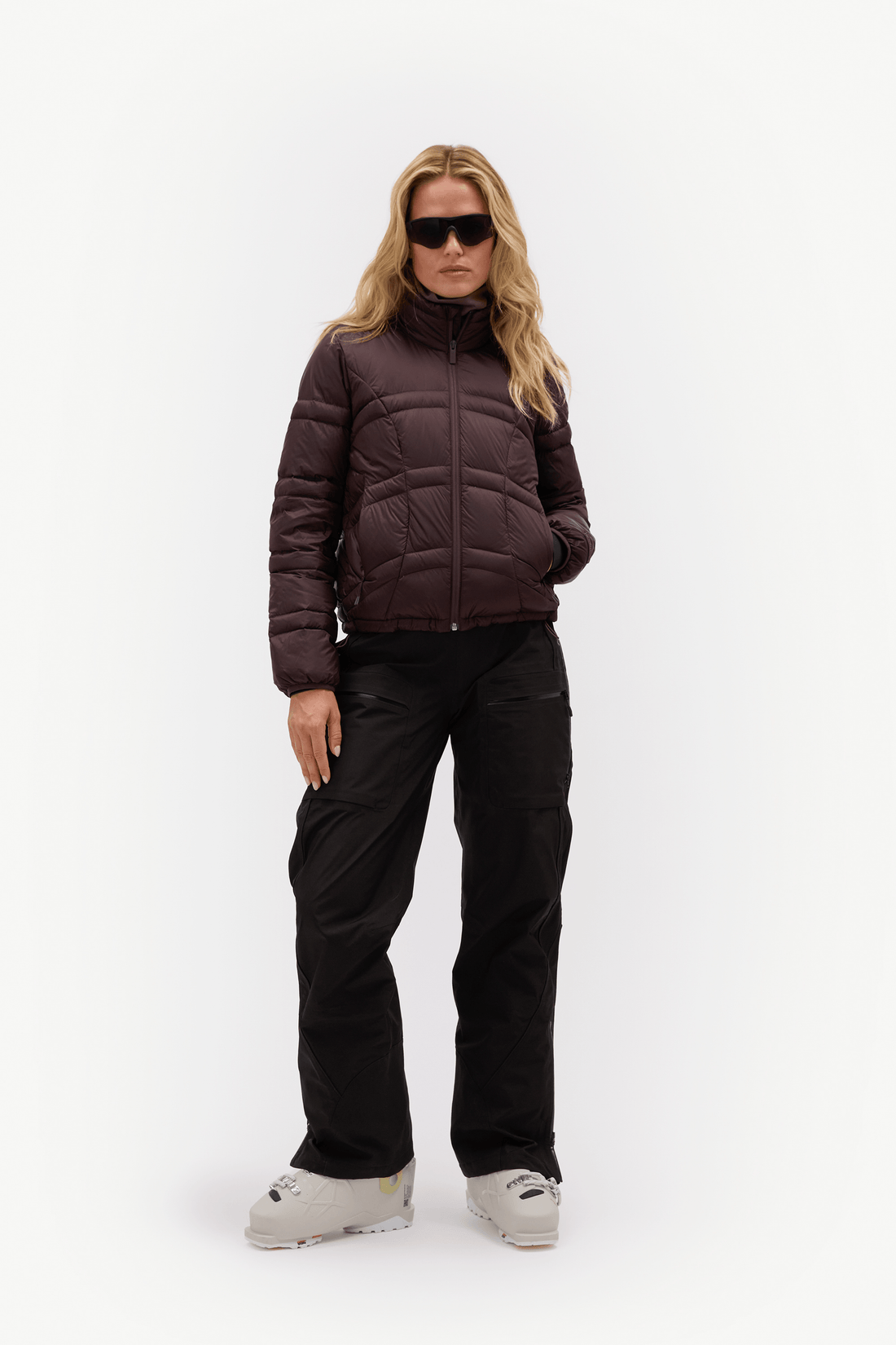 A woman modeling a dark brown puffer jacket, black snow pants, and light gray ski boots against a plain white studio background.