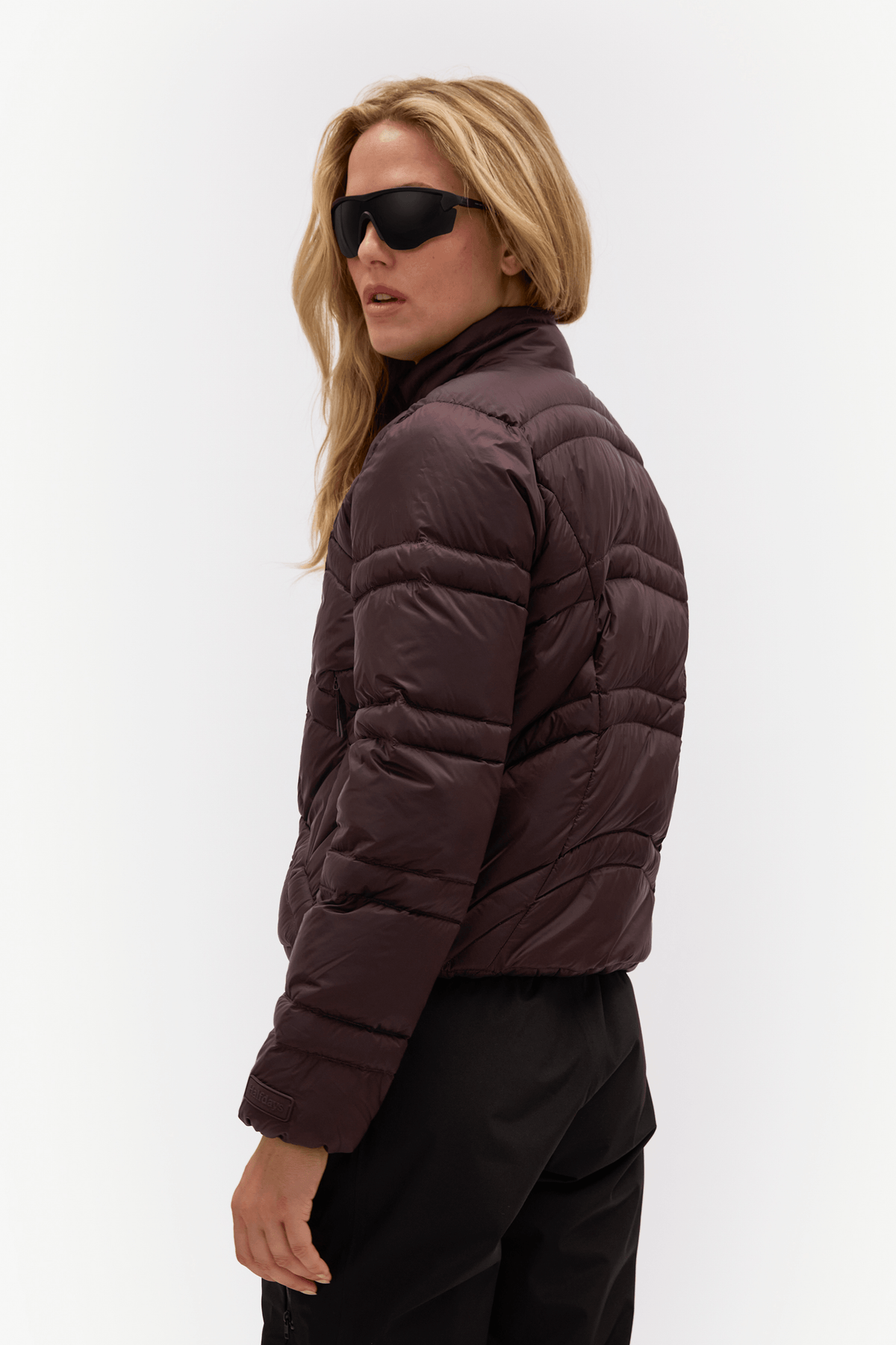 A woman wearing a dark brown cropped puffer jacket, black pants, and black sunglasses, shown from a side-back angle against a plain white studio background.