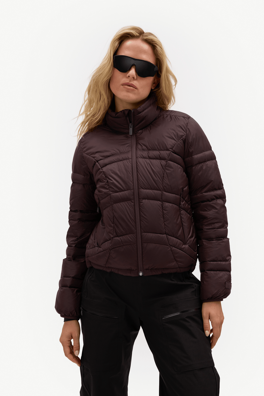 A woman modeling a dark plum purple cropped puffer jacket with a high collar and front zipper, paired with black pants, against a plain white studio background.