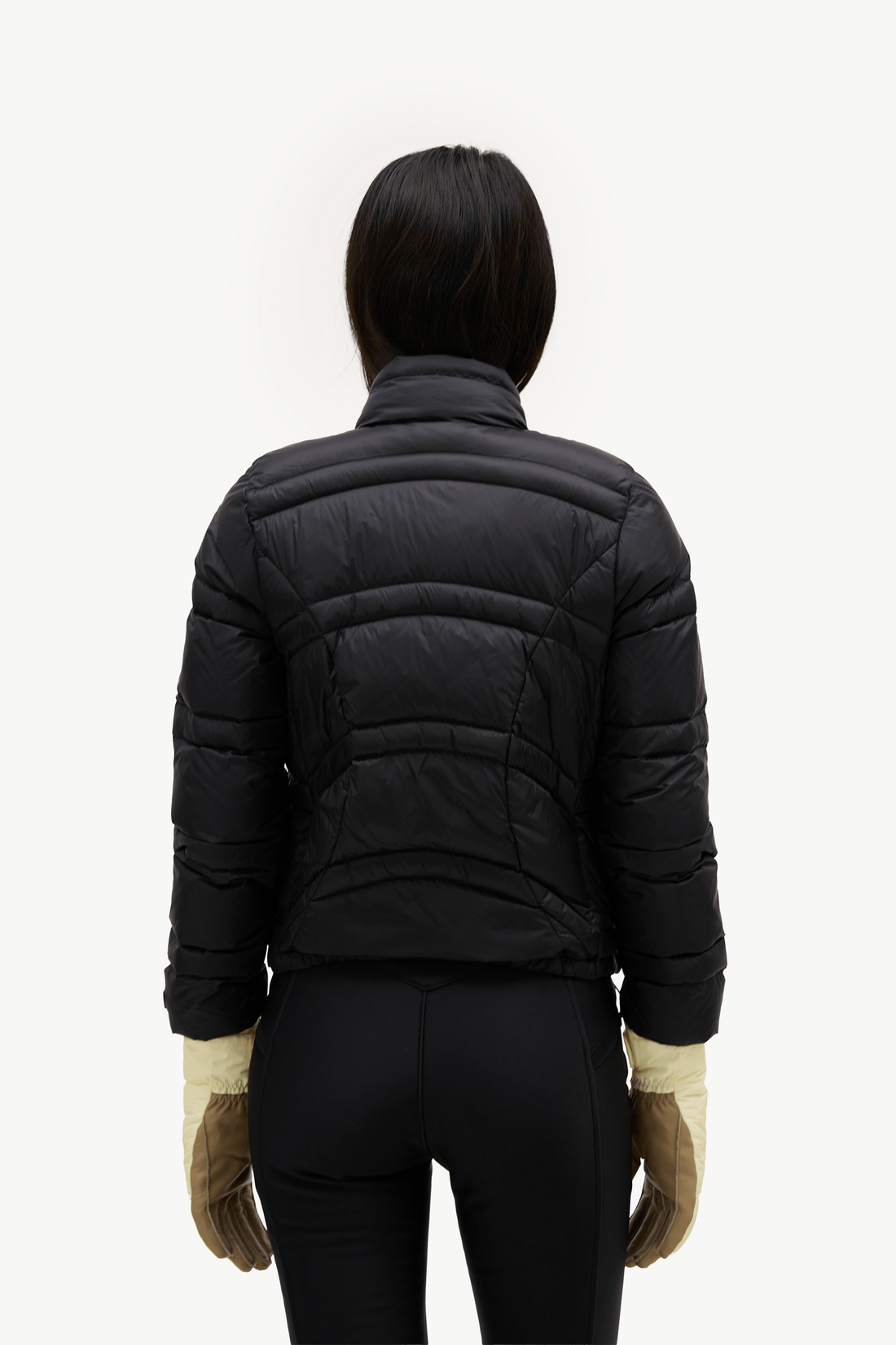 A person shown from the back wearing a black quilted puffer jacket, black pants, and tan gloves against a plain light background.
