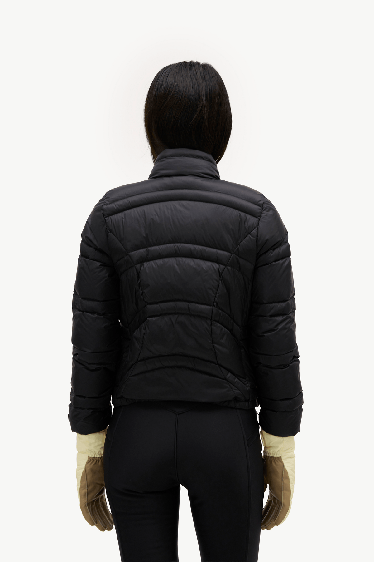 A person shown from the back wearing a black quilted puffer jacket, black pants, and tan gloves against a plain light background.