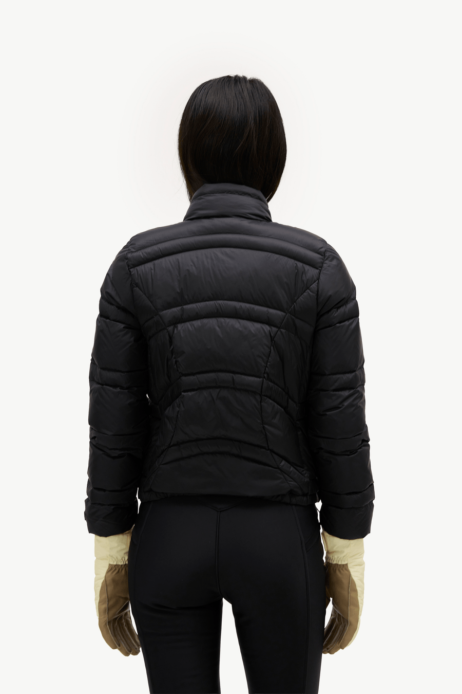 A person shown from the back wearing a black quilted puffer jacket, black pants, and tan gloves against a plain light background.