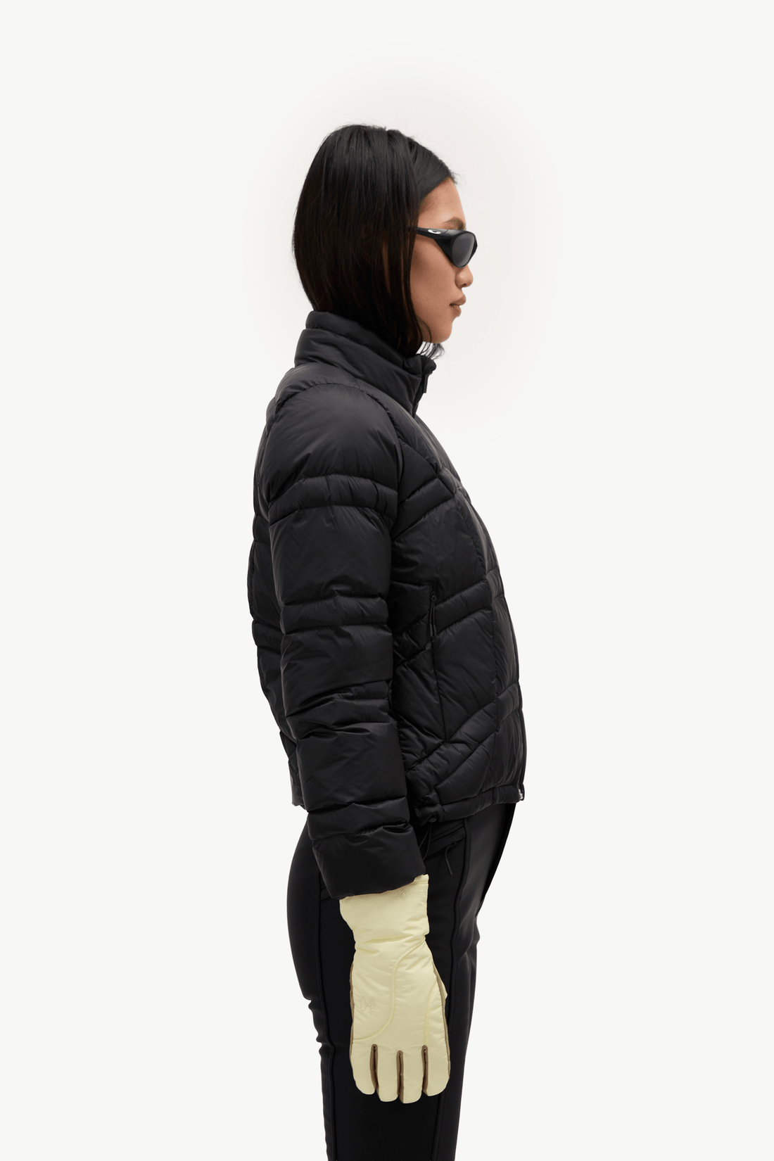 A model shown in profile wearing a black quilted puffer jacket, black pants, cream gloves, and black sunglasses against a plain white studio background.
