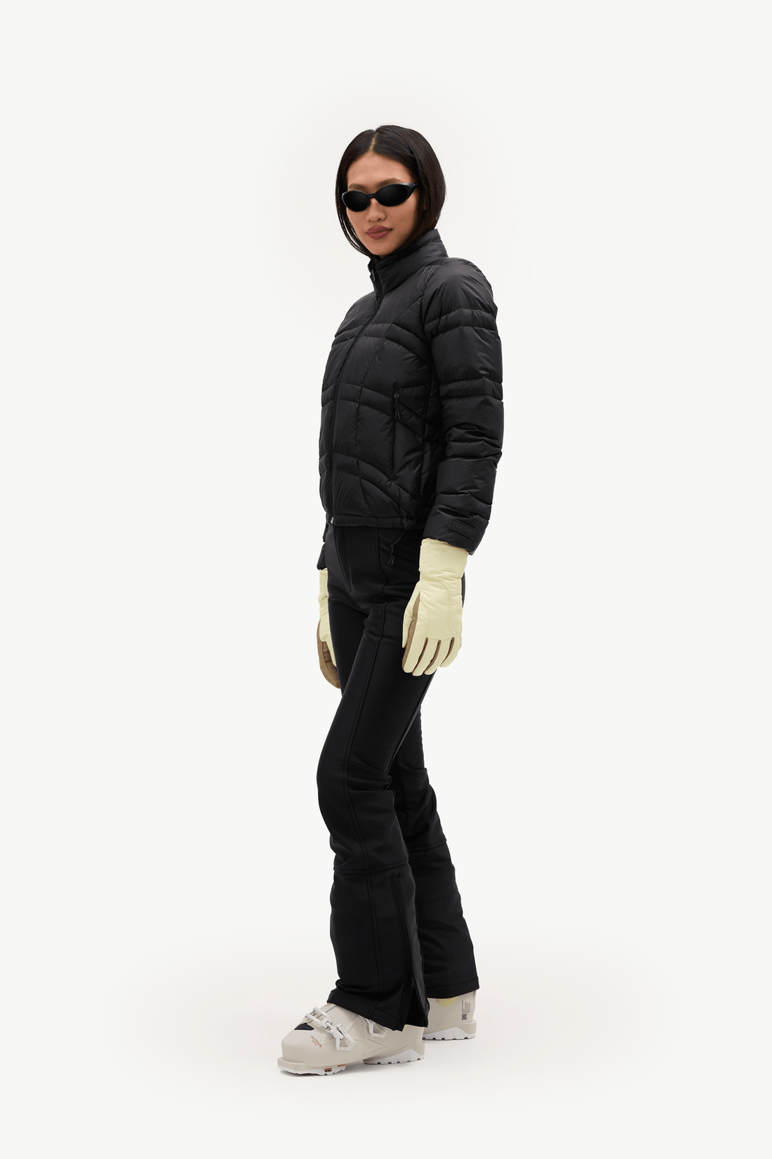 A model wearing a black puffer jacket, black ski pants, cream gloves, and light gray ski boots, standing in profile against a solid white background.