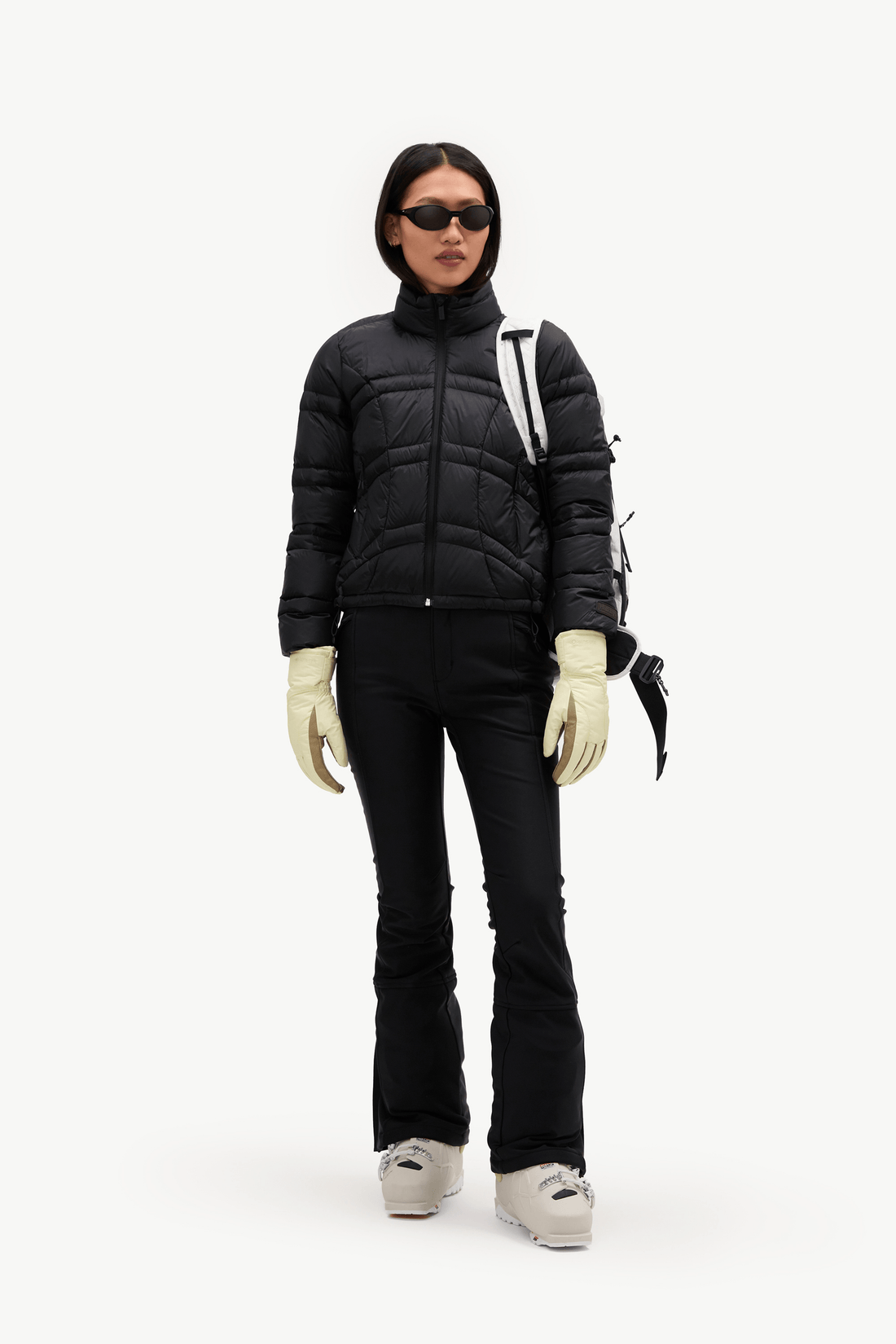 A model stands against a plain white studio background wearing a black puffer jacket, black ski pants, off-white gloves, black ski boots, and sunglasses, with a black backpack visible.