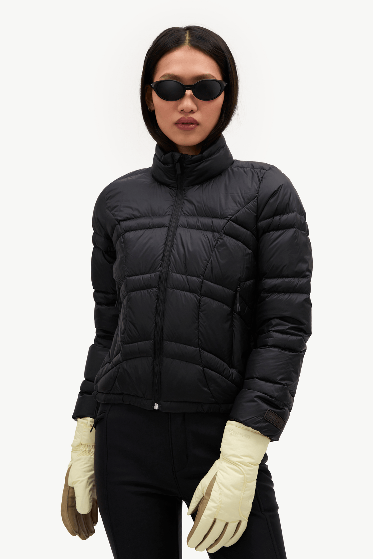 A model wearing a black puffer jacket, black pants, cream gloves, and black sunglasses, photographed against a clean white studio background.