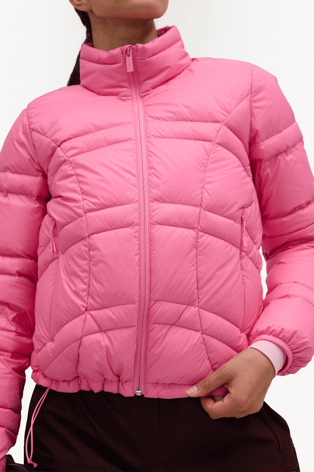 A close-up of a person wearing a bright pink quilted puffer jacket with a stand-up collar and front zipper, paired with dark brown bottoms against a plain light background.