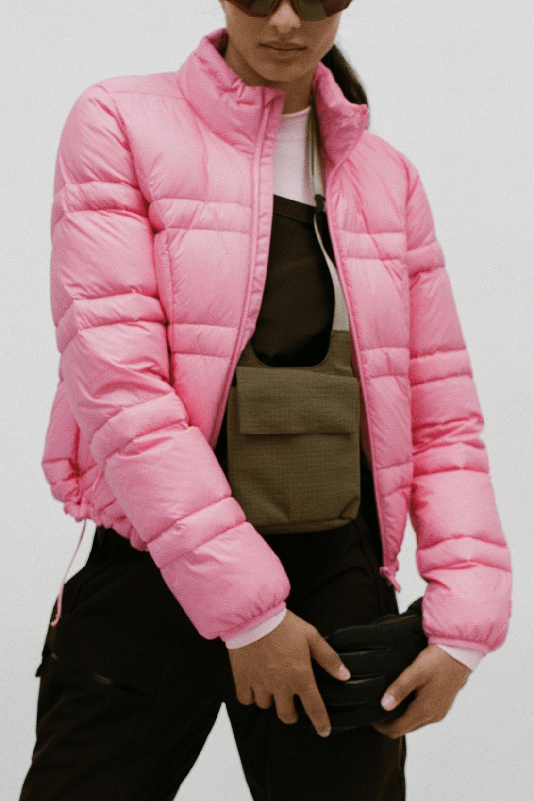 A model wearing a bright pink puffer jacket over a brown vest, black pants, and sunglasses, holding a black clutch bag against a plain light gray background.