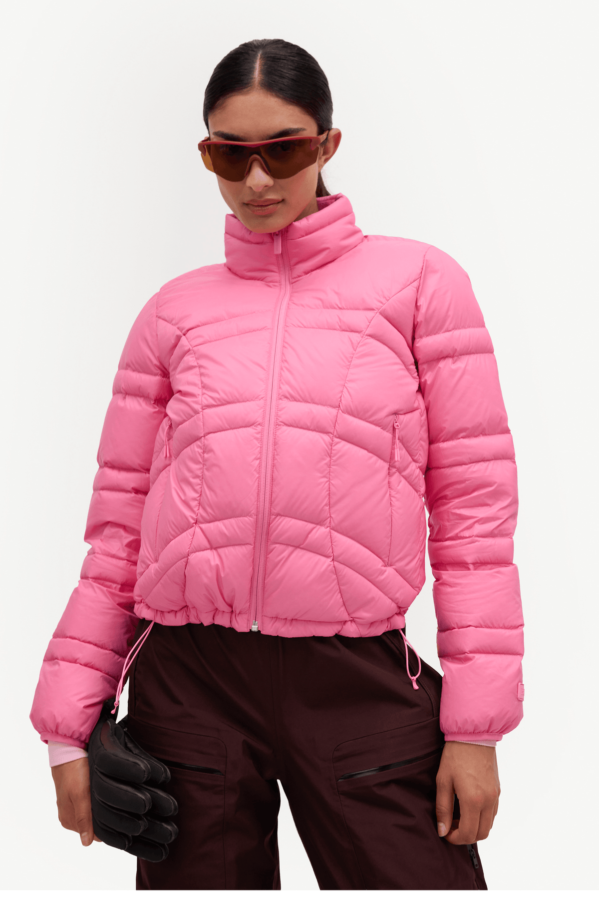 A model wearing a bright pink cropped puffer jacket with a high collar, paired with dark brown pants, black gloves, and sunglasses, against a plain light gray background.