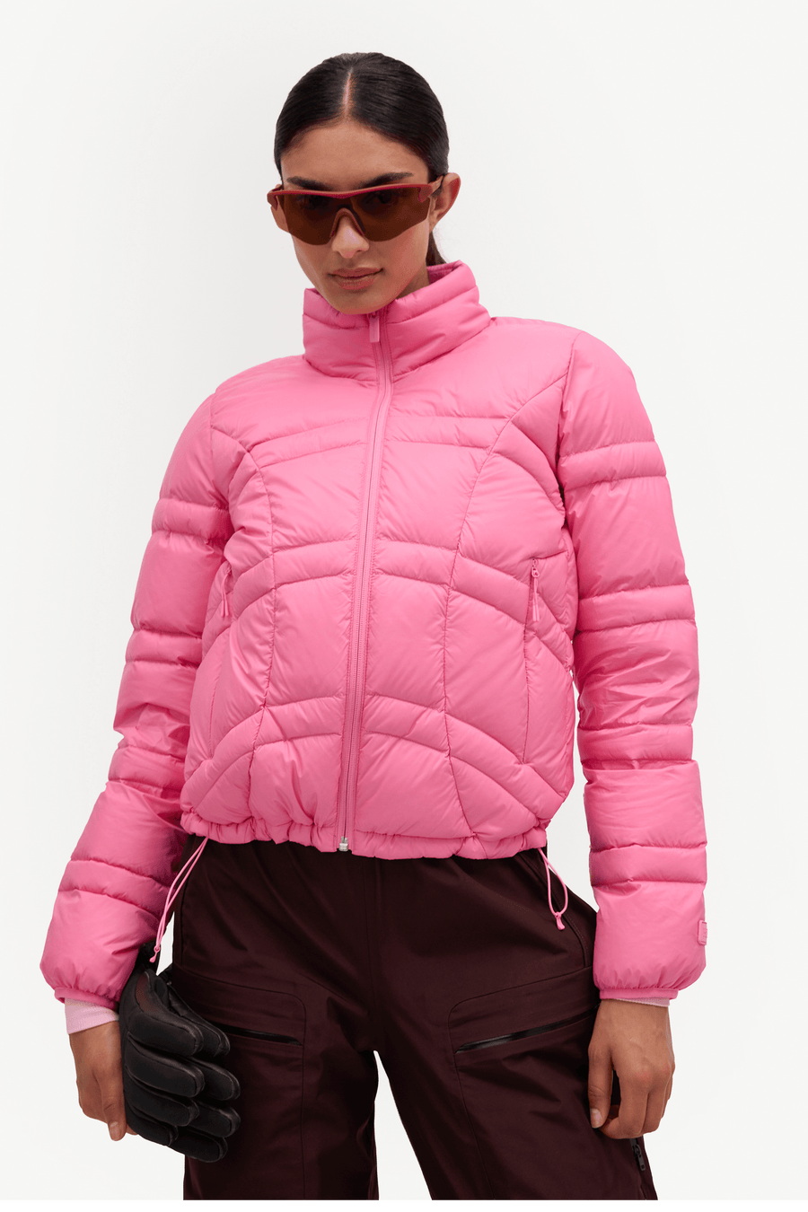 A model wearing a bright pink cropped puffer jacket with a high collar, paired with dark brown pants, black gloves, and sunglasses, against a plain light gray background.