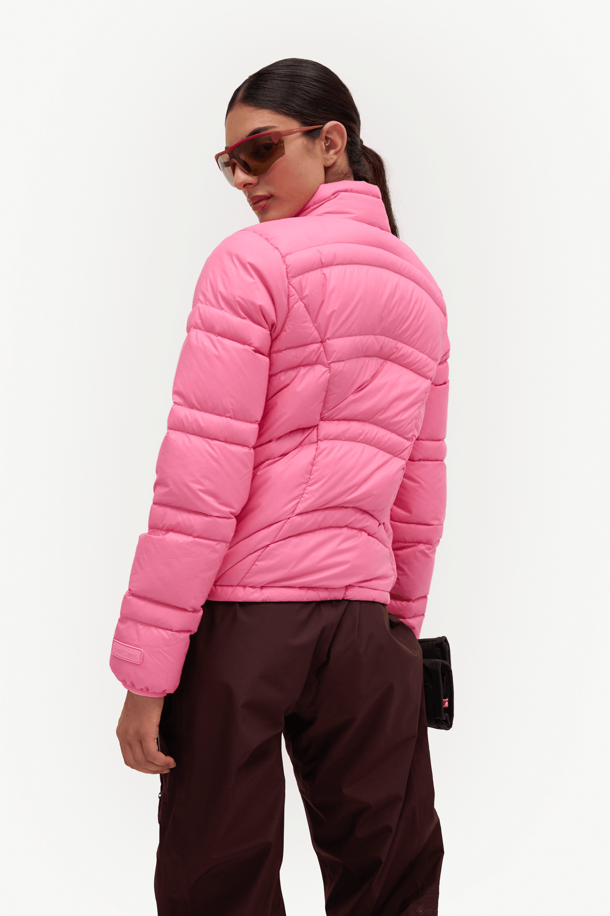 A woman shown from the back wearing a bright pink quilted puffer jacket and dark brown pants against a clean white studio background.