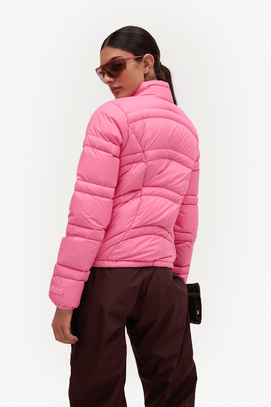 A woman shown from the back wearing a bright pink quilted puffer jacket and dark brown pants against a clean white studio background.