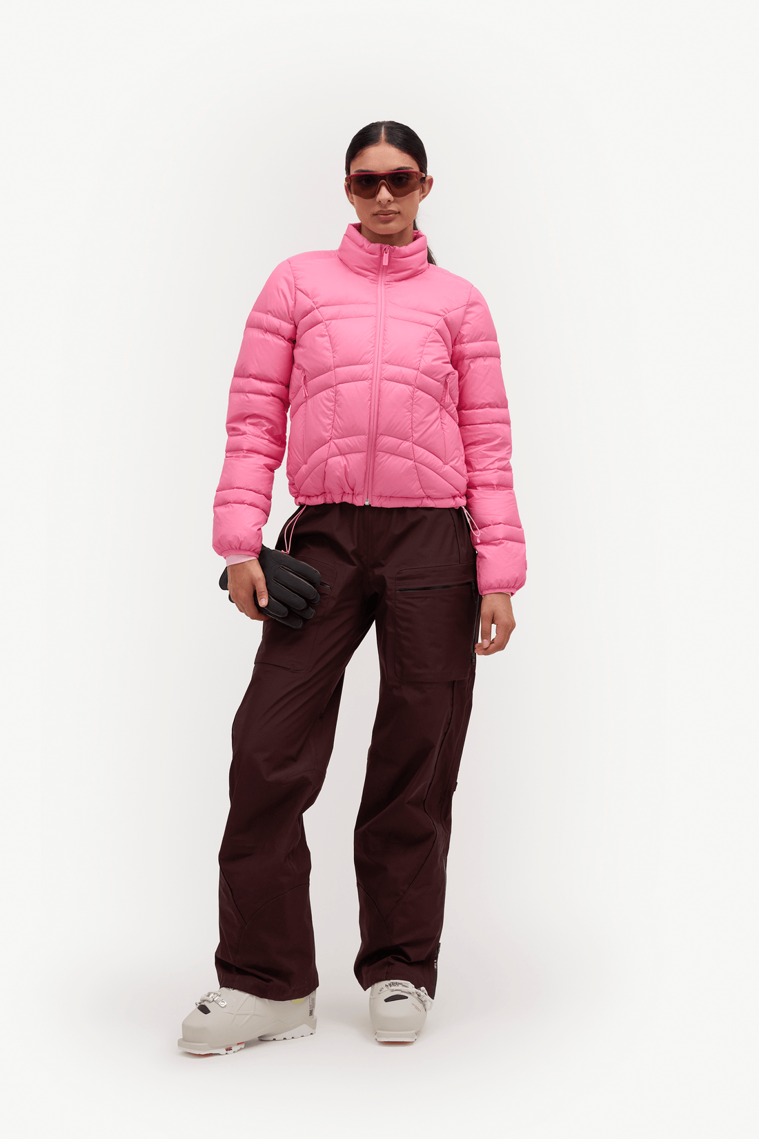 A model wearing a bright pink puffer jacket, dark brown snow pants, black gloves, and light gray snow boots, standing against a plain white studio background.