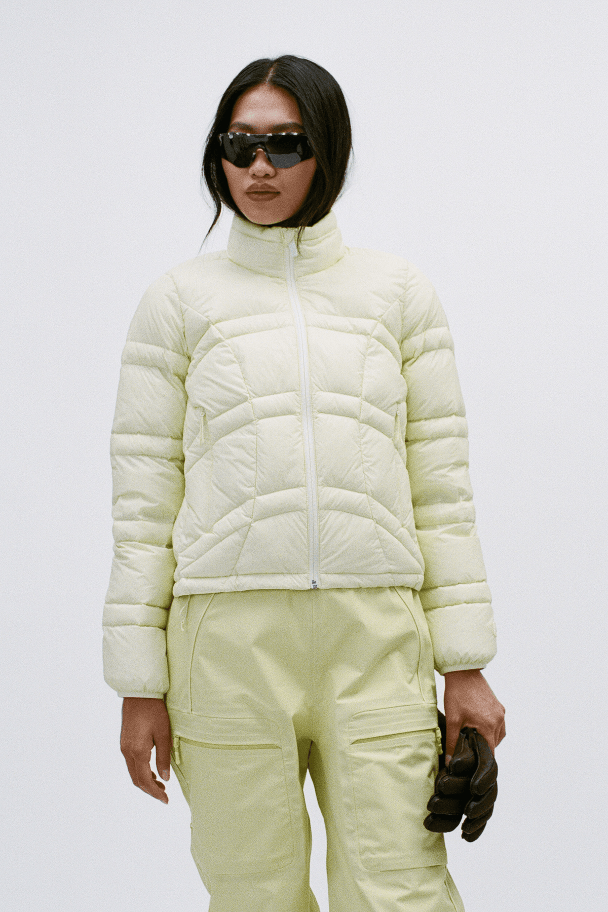 A model wearing a pale yellow quilted puffer jacket, matching light yellow pants, black sunglasses, and holding a black glove against a plain light gray studio background.