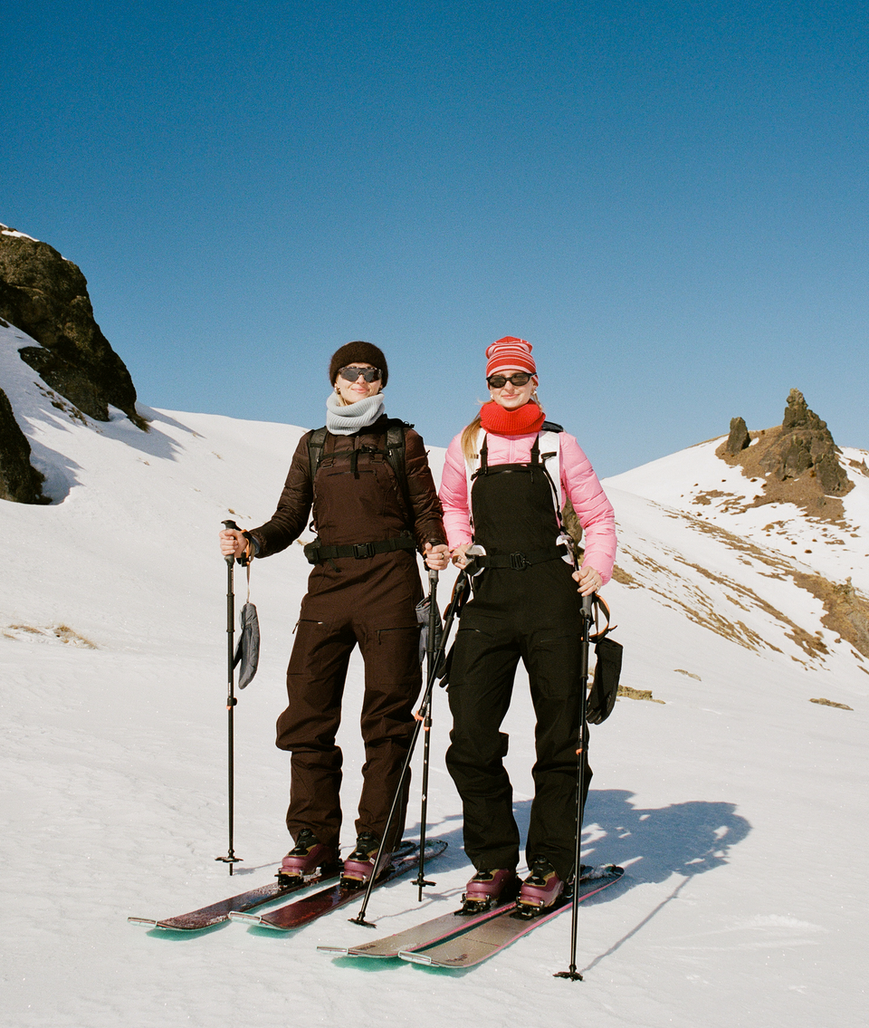 Two skiers standing on a snowy mountain slope under a clear blue sky, wearing winter sports gear and holding ski poles. One skier is dressed in a dark brown outfit with a gray beanie and face covering, and the other in a pink jacket with black overalls, a red beanie, and a red face covering.
