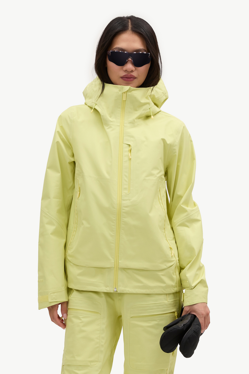 A model wearing a light yellow-green ski jacket and matching pants, holding black gloves against a plain white studio background.