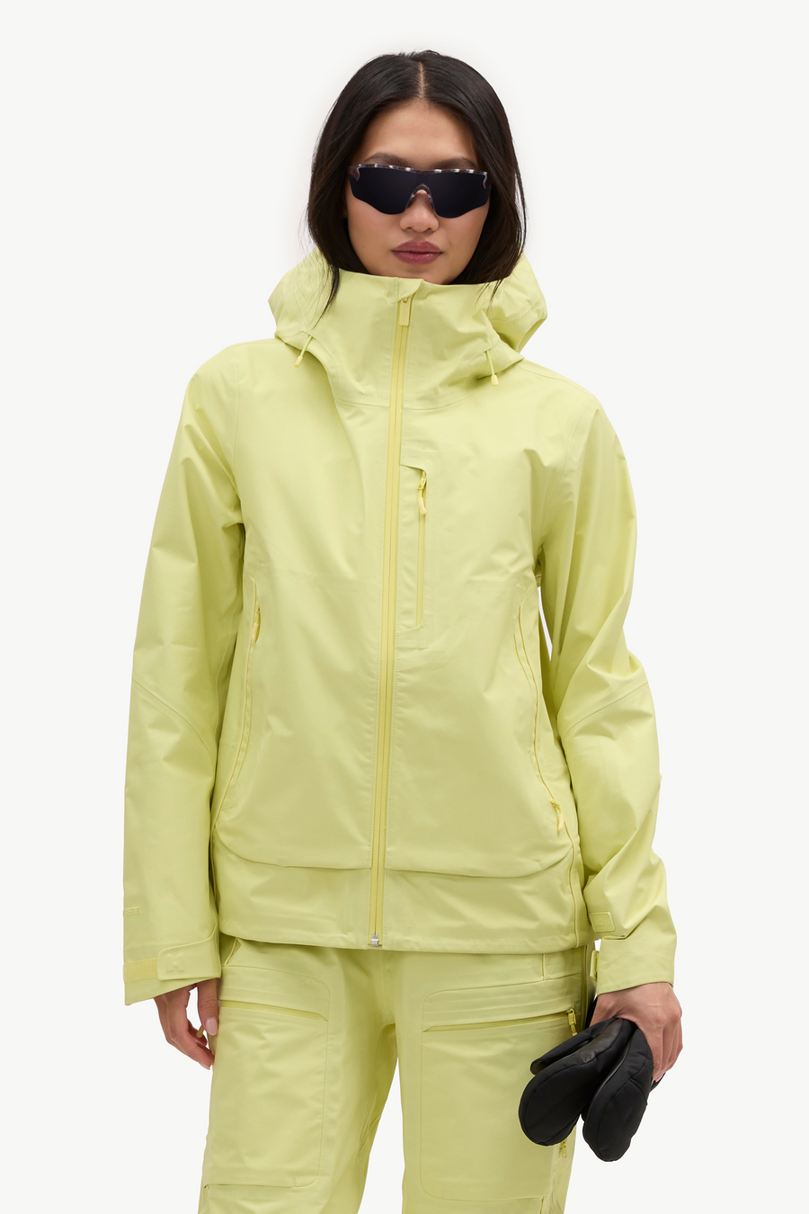 A model wearing a light yellow-green ski jacket and matching pants, holding black gloves against a plain white studio background.