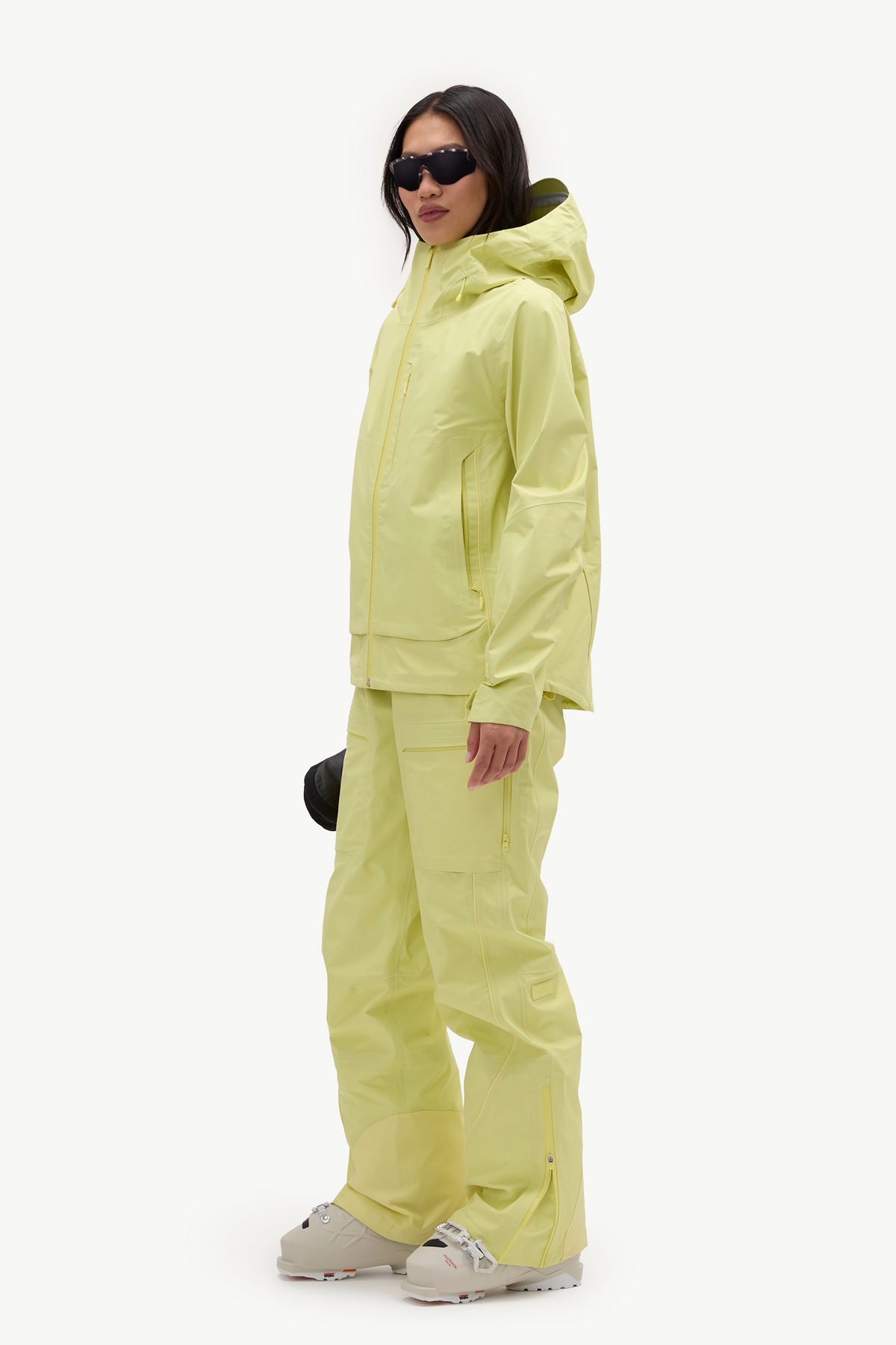 A model wearing a matching light yellow-green hooded jacket and pants set, styled with beige snowboard boots and sunglasses, set against a plain white studio background.