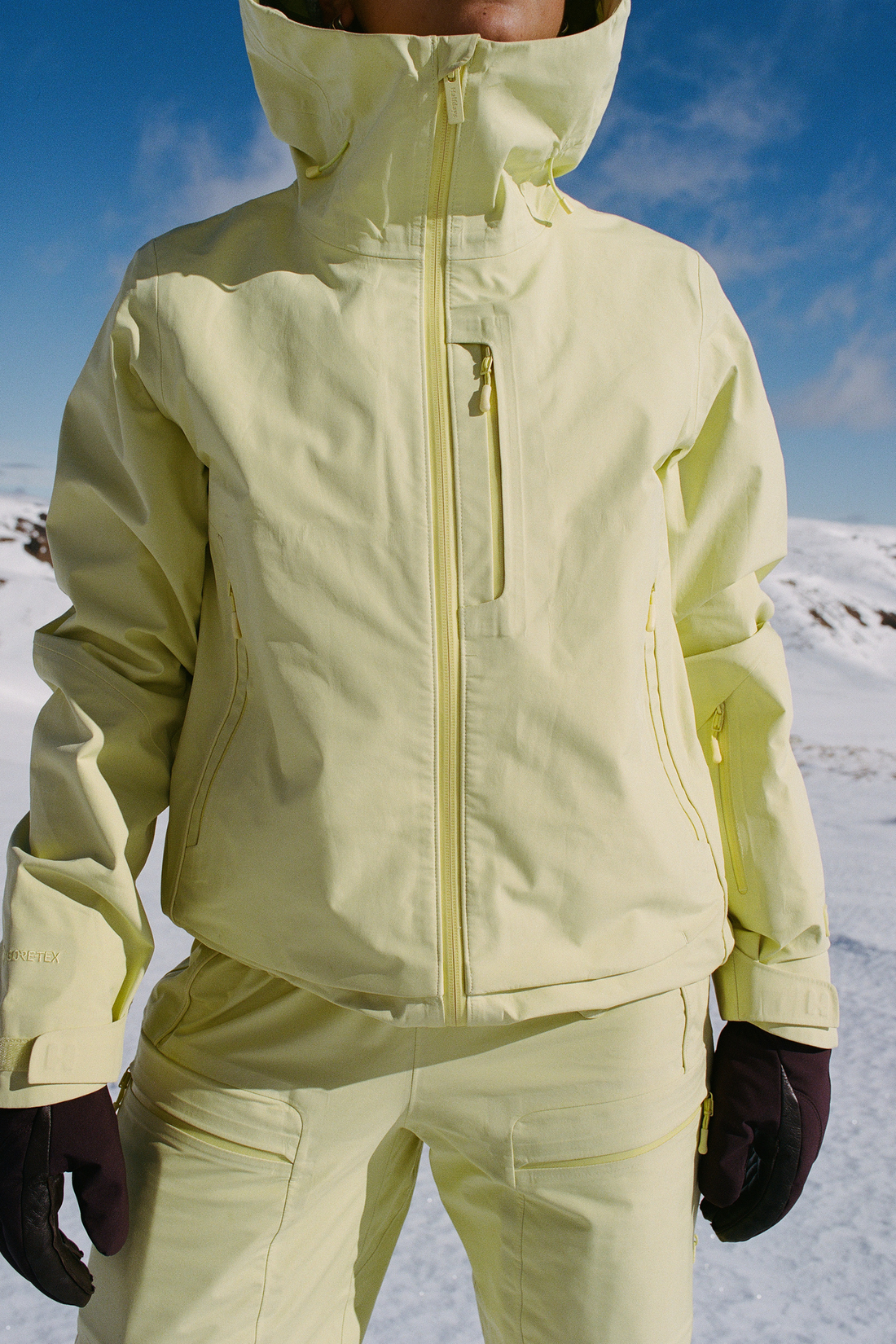 A person wearing a light yellow ski jacket and matching pants in a snowy outdoor setting under a blue sky.