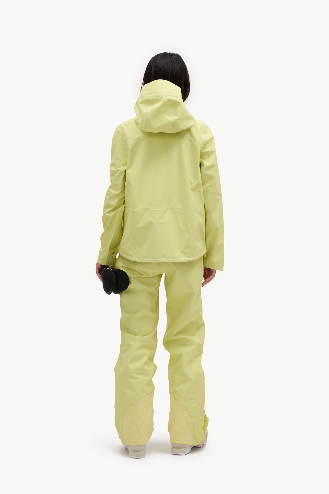 A model viewed from the back wearing a light yellow-green hooded jacket and matching pants, holding a black beanie against a plain white studio background.
