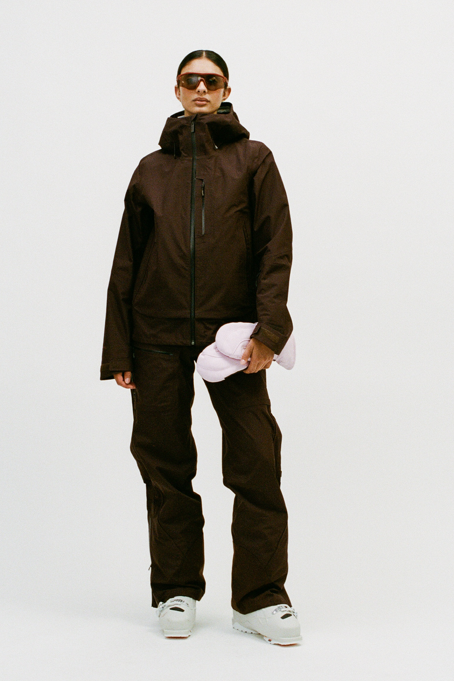 A model wearing a dark brown ski jacket and matching pants, holding a light pink ski helmet, standing against a plain white studio background.