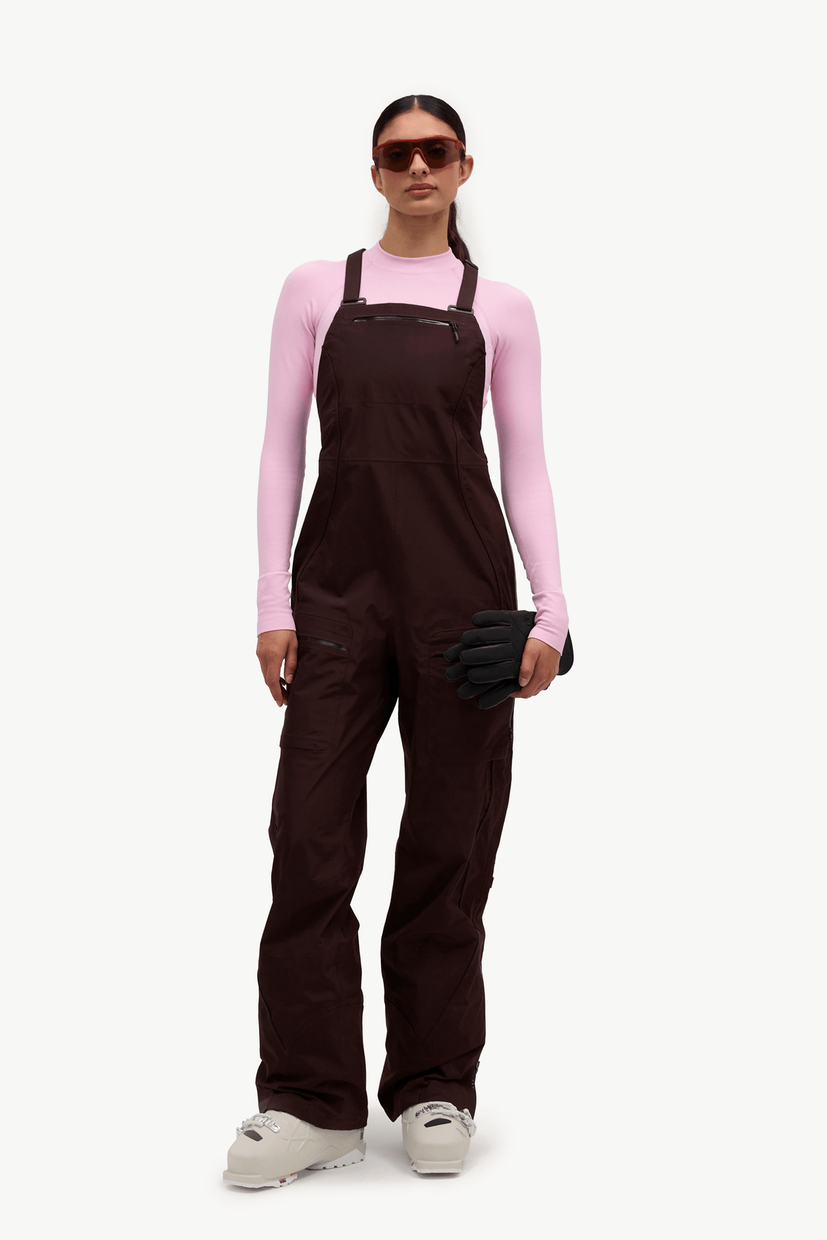 A model wearing dark brown snow bib overalls over a light pink long-sleeve shirt, holding a black glove, and wearing white snowboard boots, on a solid white background.