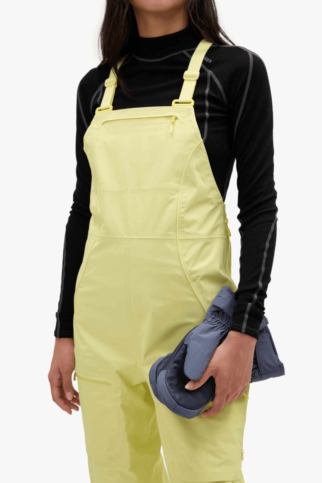 A model wearing light yellow bib snow pants over a black long-sleeve top, holding a pair of dark blue insulated gloves against a clean white background.
