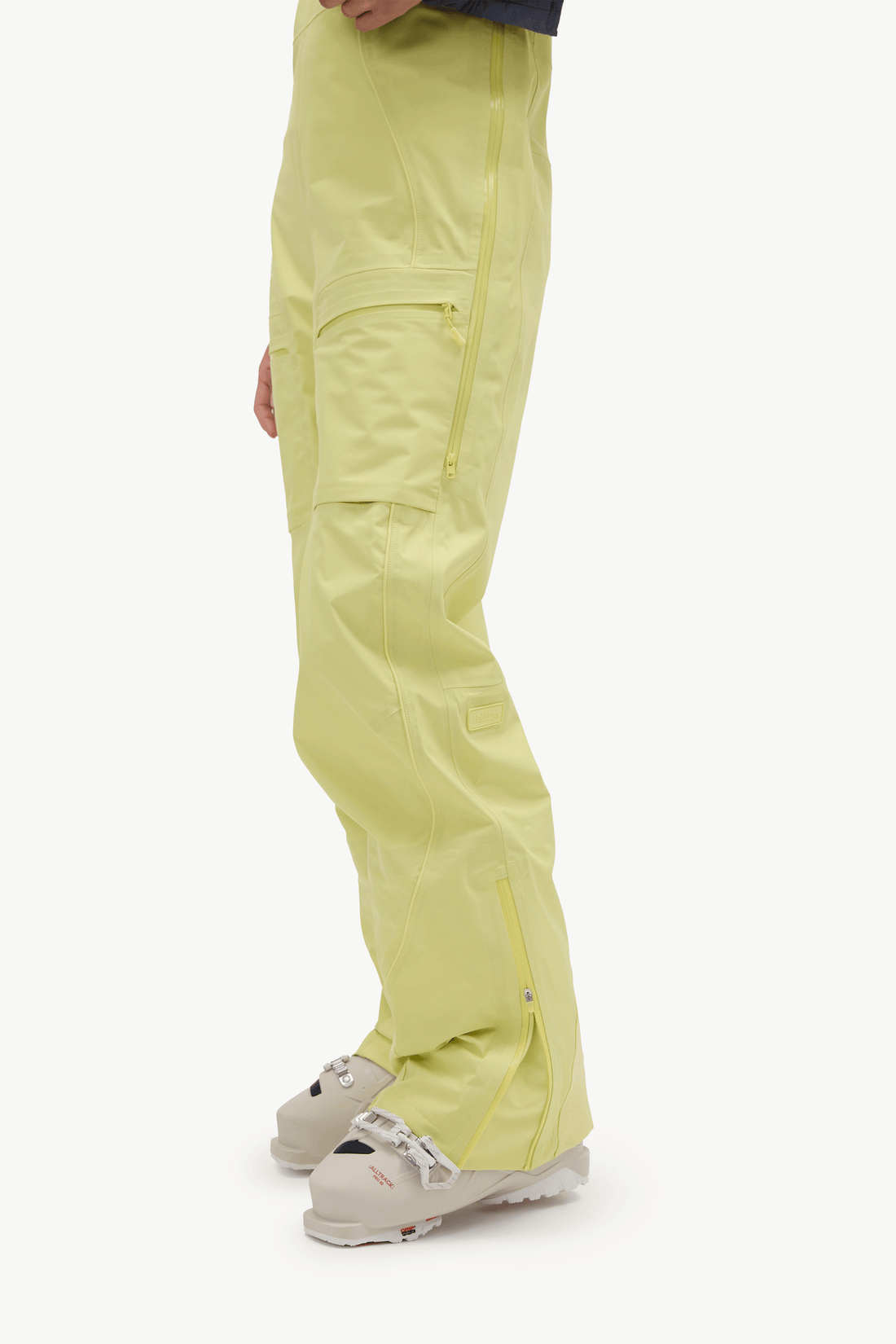 A side profile view of a person wearing light yellow-green ski pants with zippered cargo pockets and light-colored ski boots, set against a plain white studio background.