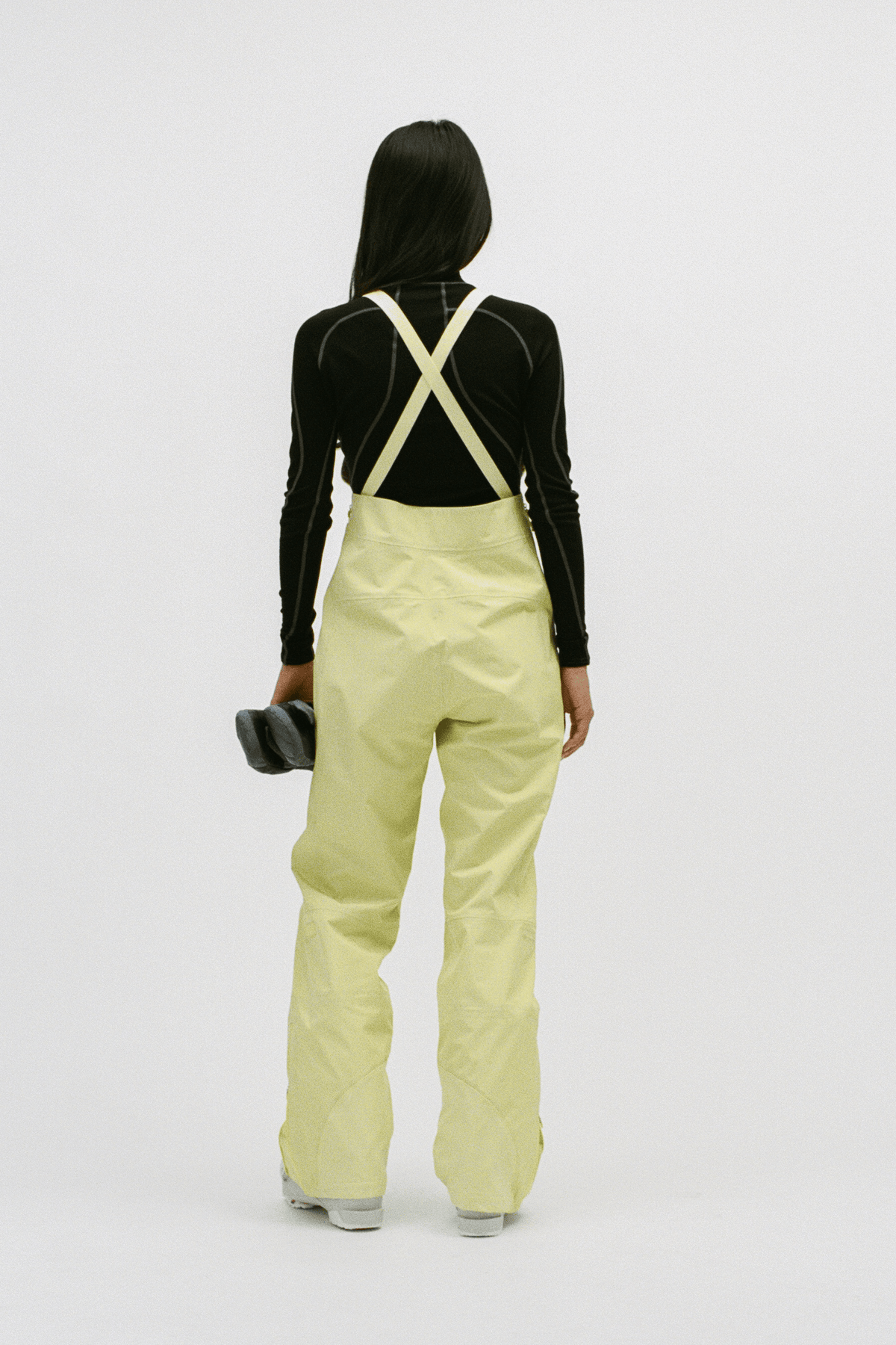 A model shown from the back wearing light yellow snow pants with cream-colored suspenders over a black long-sleeve top, holding a black ski boot against a plain white studio background.